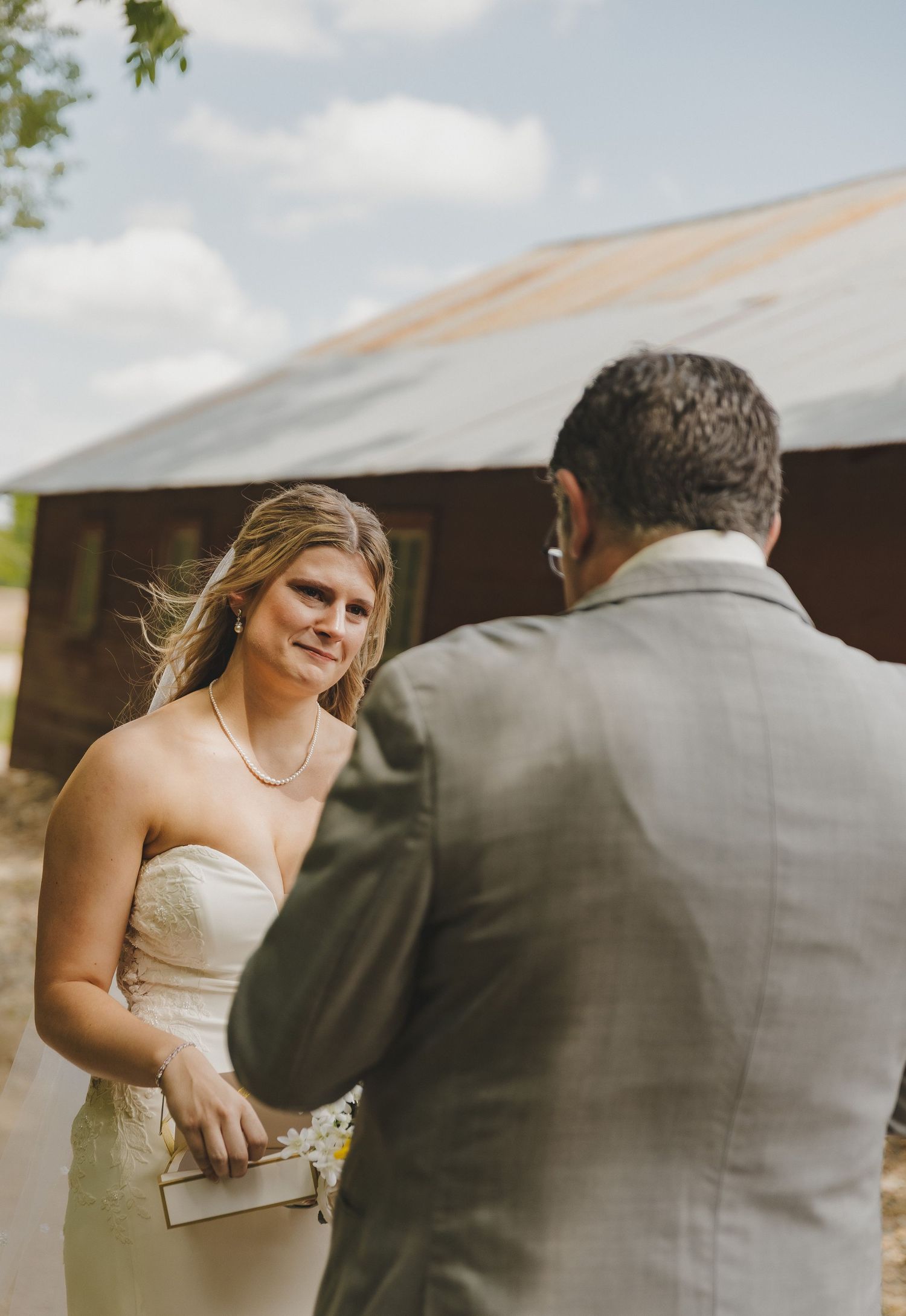 Bride in strapless white gown reads vows to groom in gray suit during outdoor rustic barn wedding ceremony.