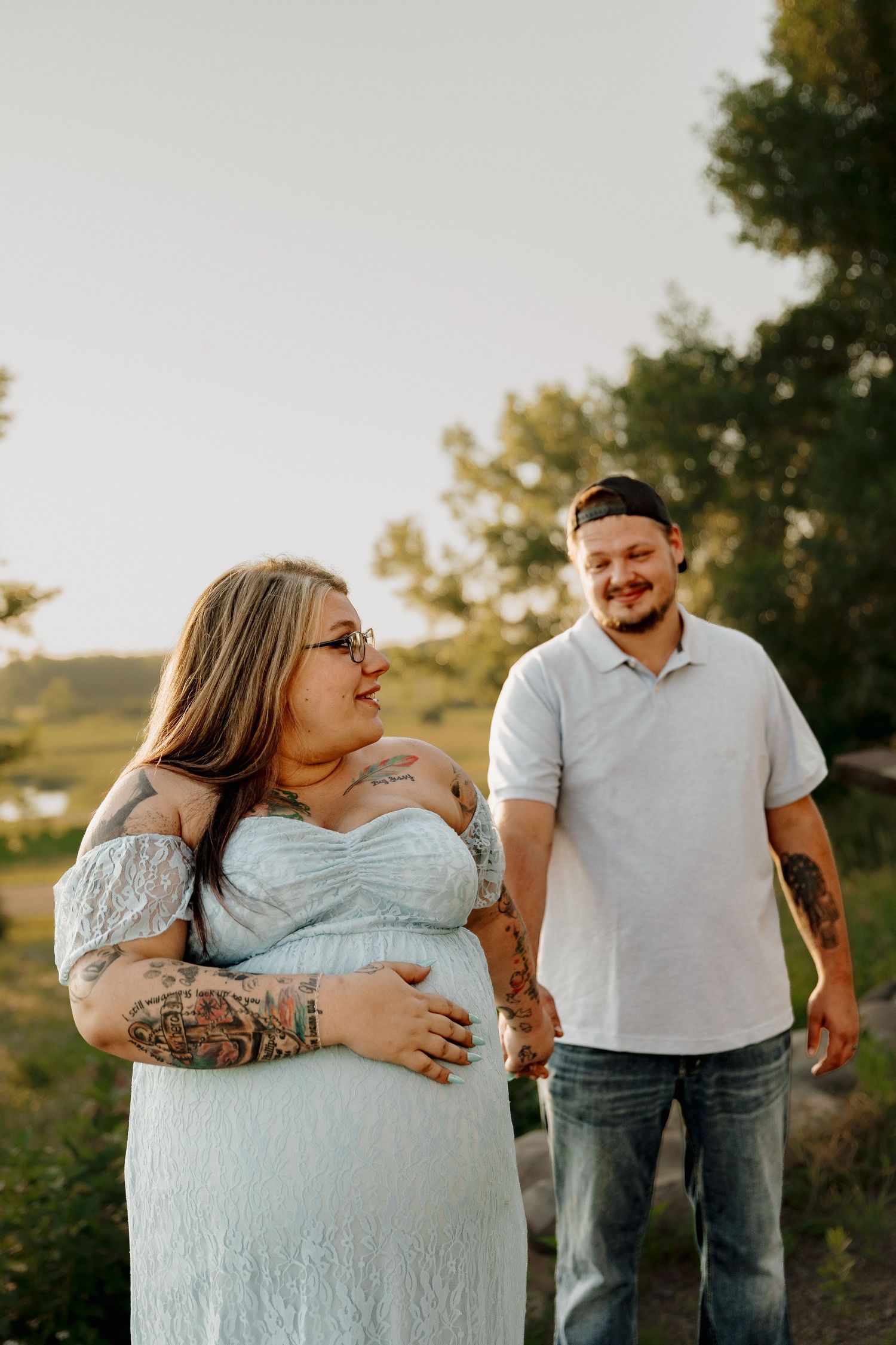 Pregnant woman in light blue dress holds baby bump while tattooed man in white polo smiles at her outdoors.