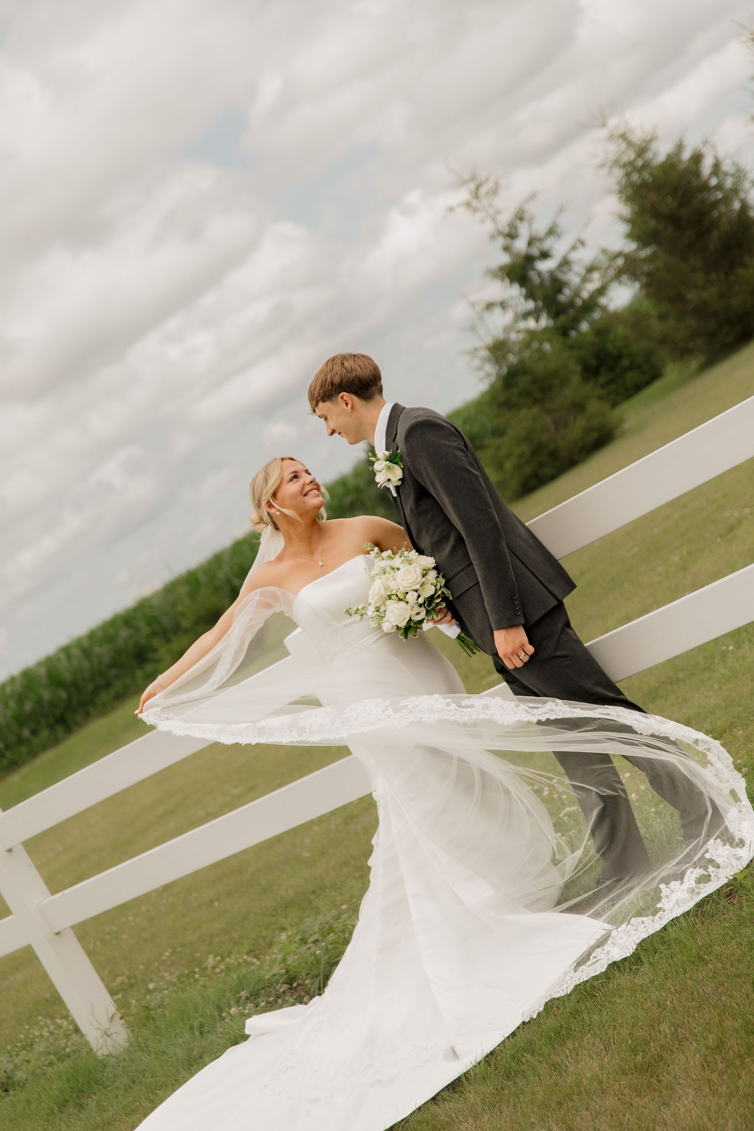 Bride in flowing white gown and veil stands with groom in gray suit near white fence on green countryside.