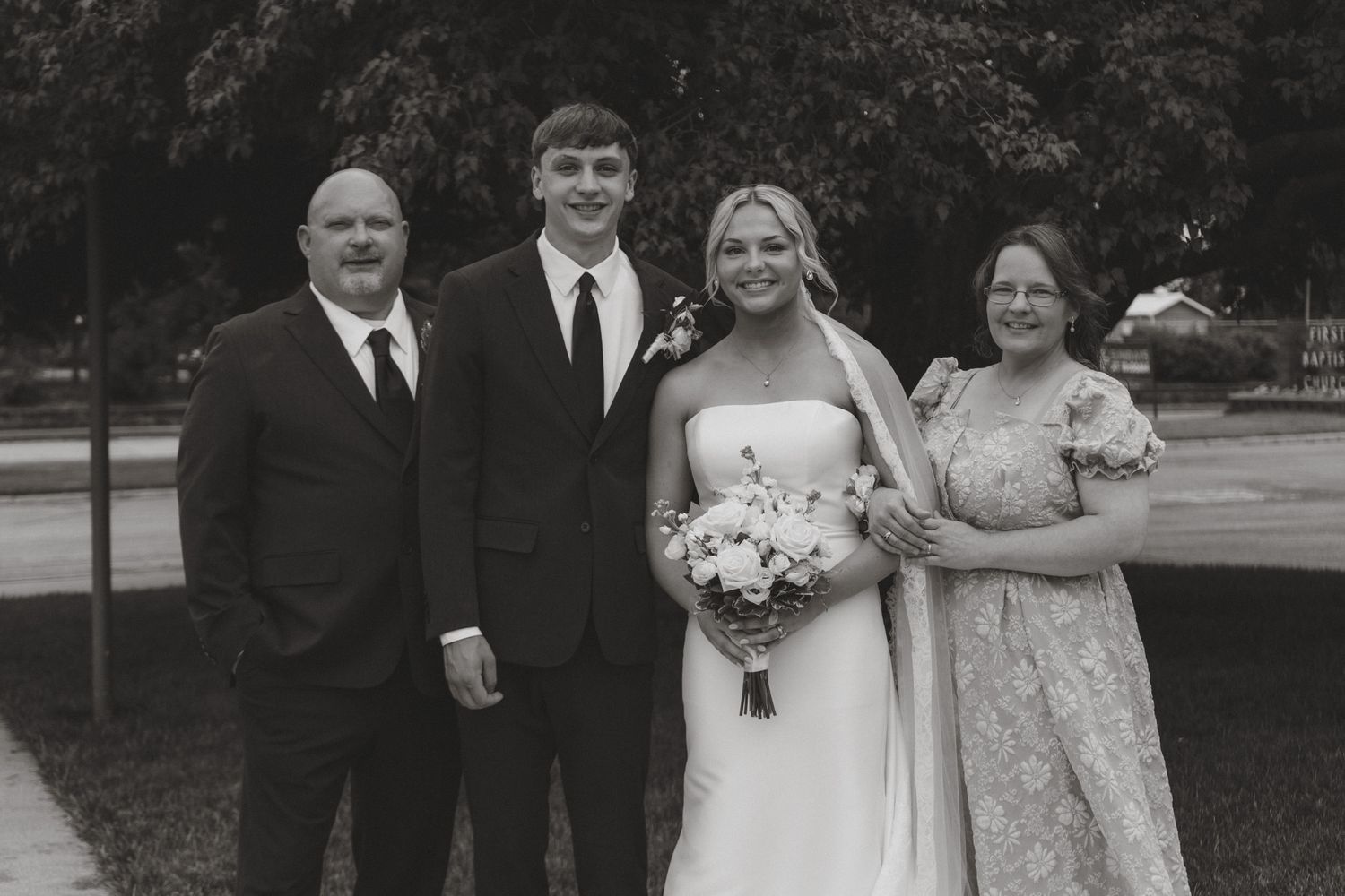 Black and white wedding photo of bride holding bouquet posing with groom and two family members outdoors.