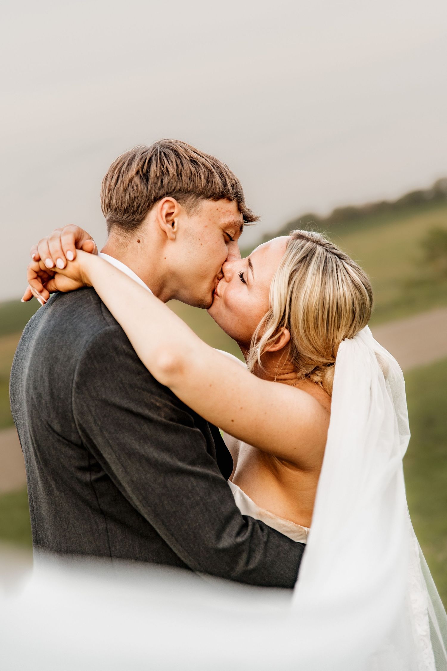 Bride and groom sharing a romantic kiss outdoors with a scenic green hillside backdrop on their wedding day.