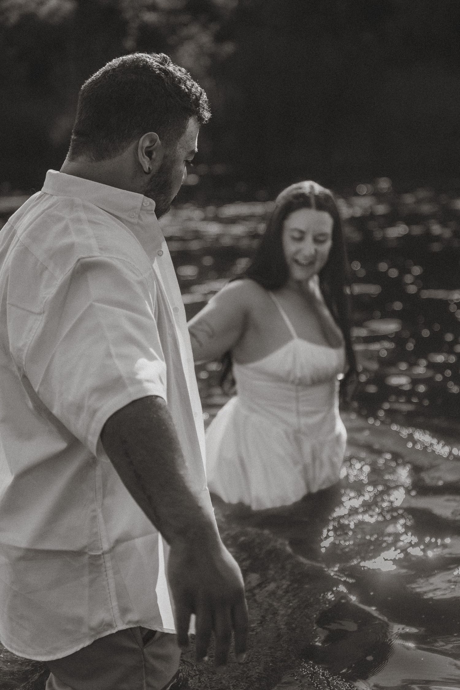 Black and white photo of a couple wading in water, man in white shirt leads woman in white dress through a sunlit river.