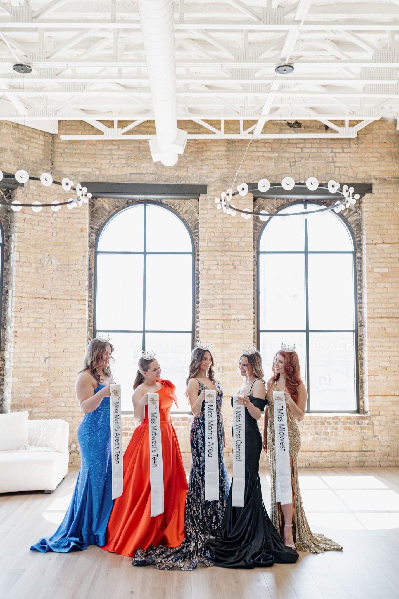 Five pageant queens in colorful gowns display sashes in a bright brick loft venue.