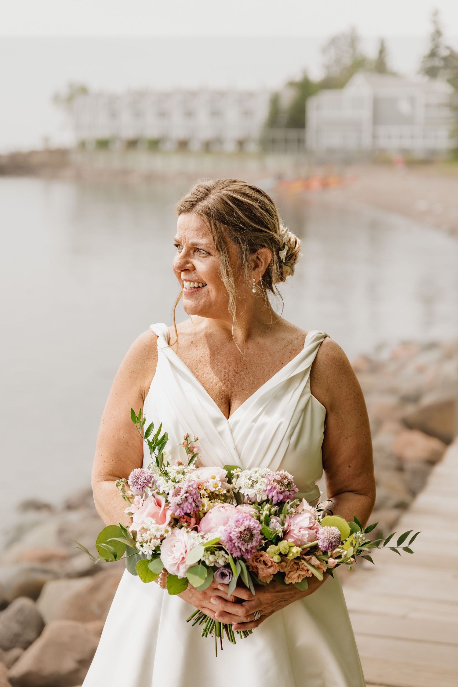Smiling bride in white gown holds pink floral bouquet on rocky waterfront with building in background.