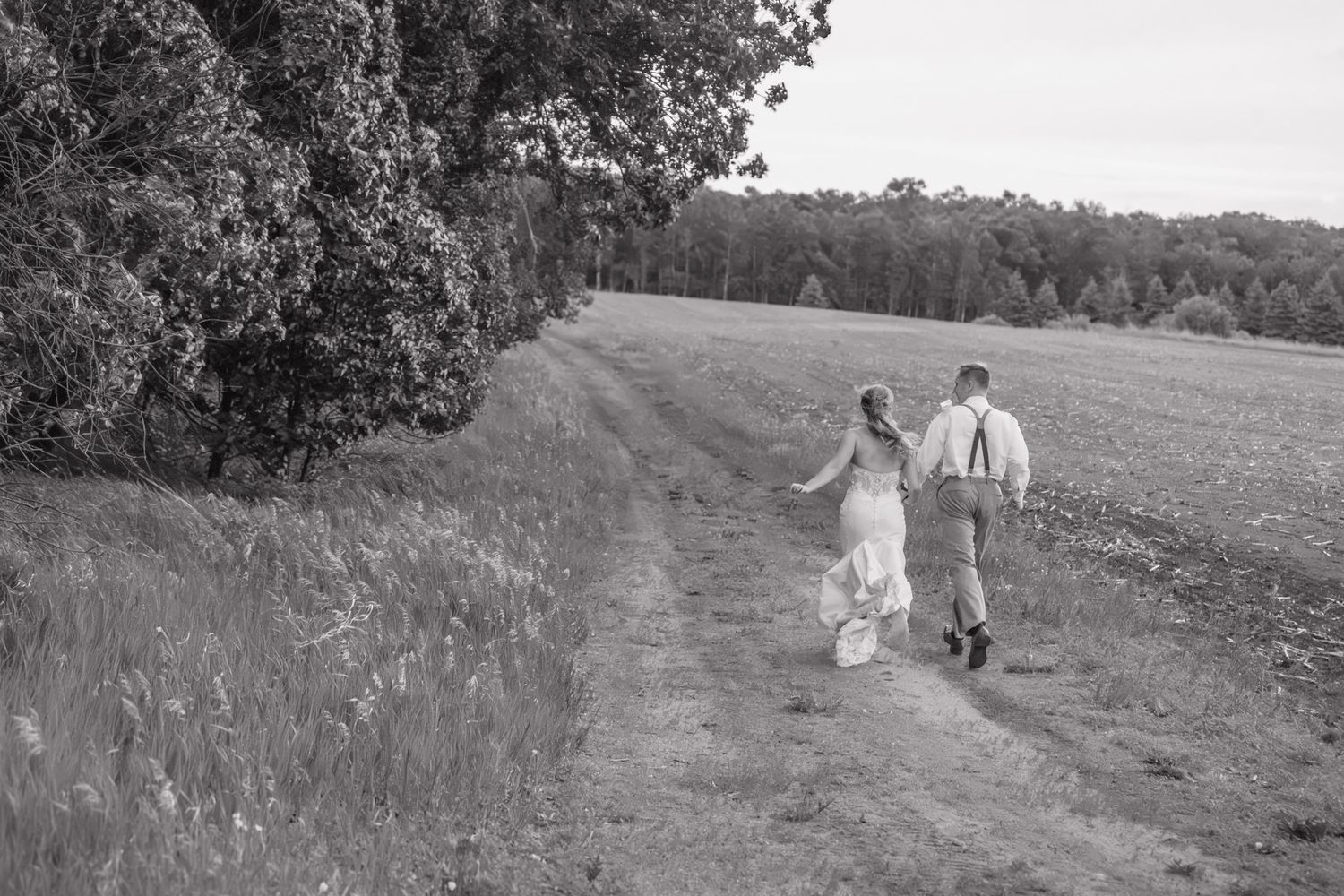 Bride and groom walk hand-in-hand along a scenic rural path on their wedding day.