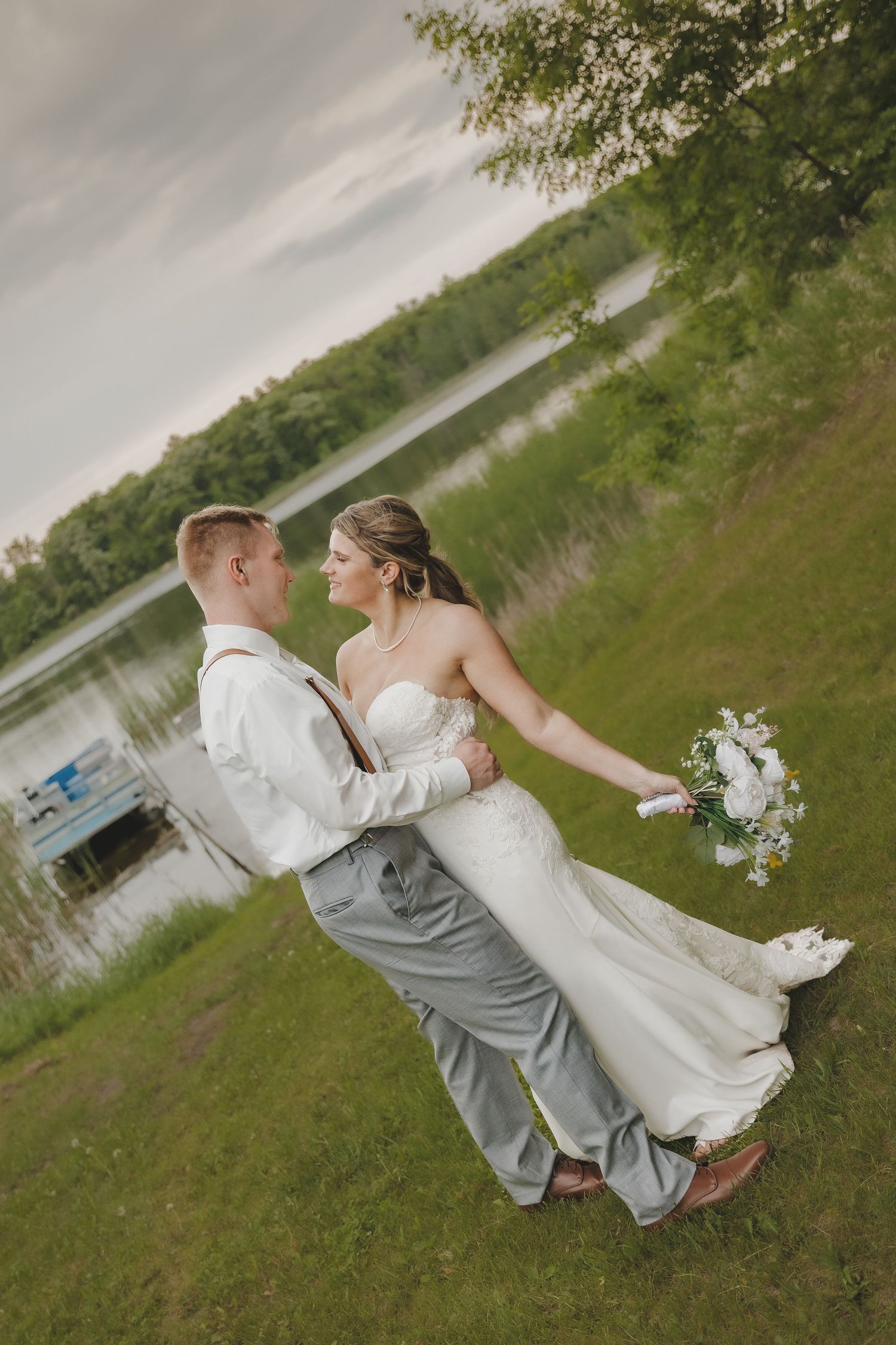 Groom lifts smiling bride in strapless gown near a serene lakeside setting at golden hour.