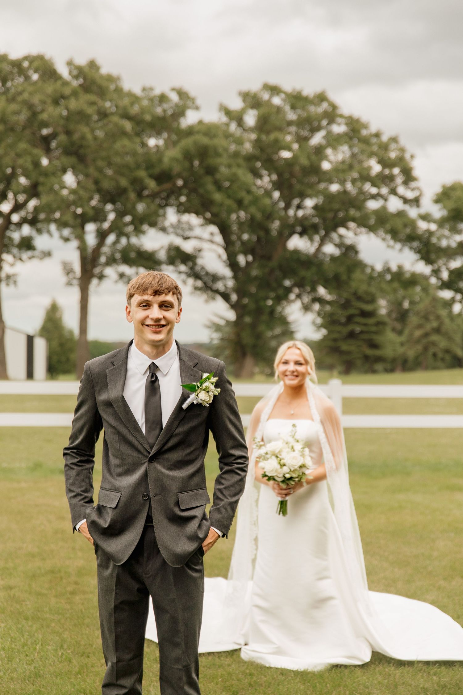 Smiling groom in gray suit stands before bride in white wedding dress holding bouquet on green lawn.