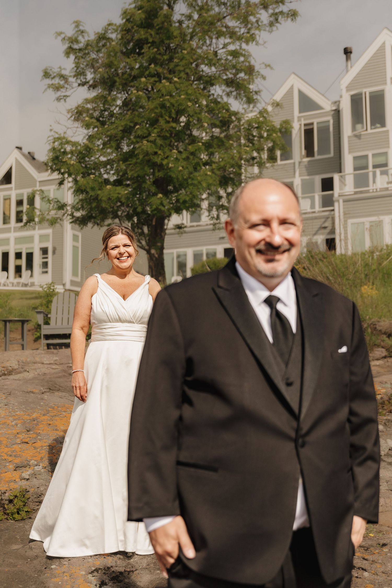 A smiling bride in a white gown stands behind a man in a black suit during a first look moment at a waterfront venue.