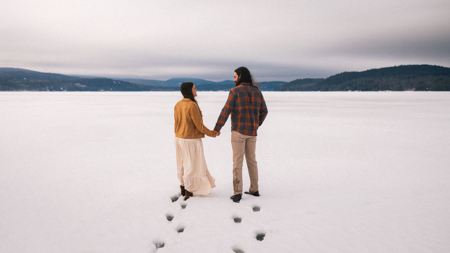 Couple holding hands standing on a frozen snow-covered lake in winter, with mountains in the background.