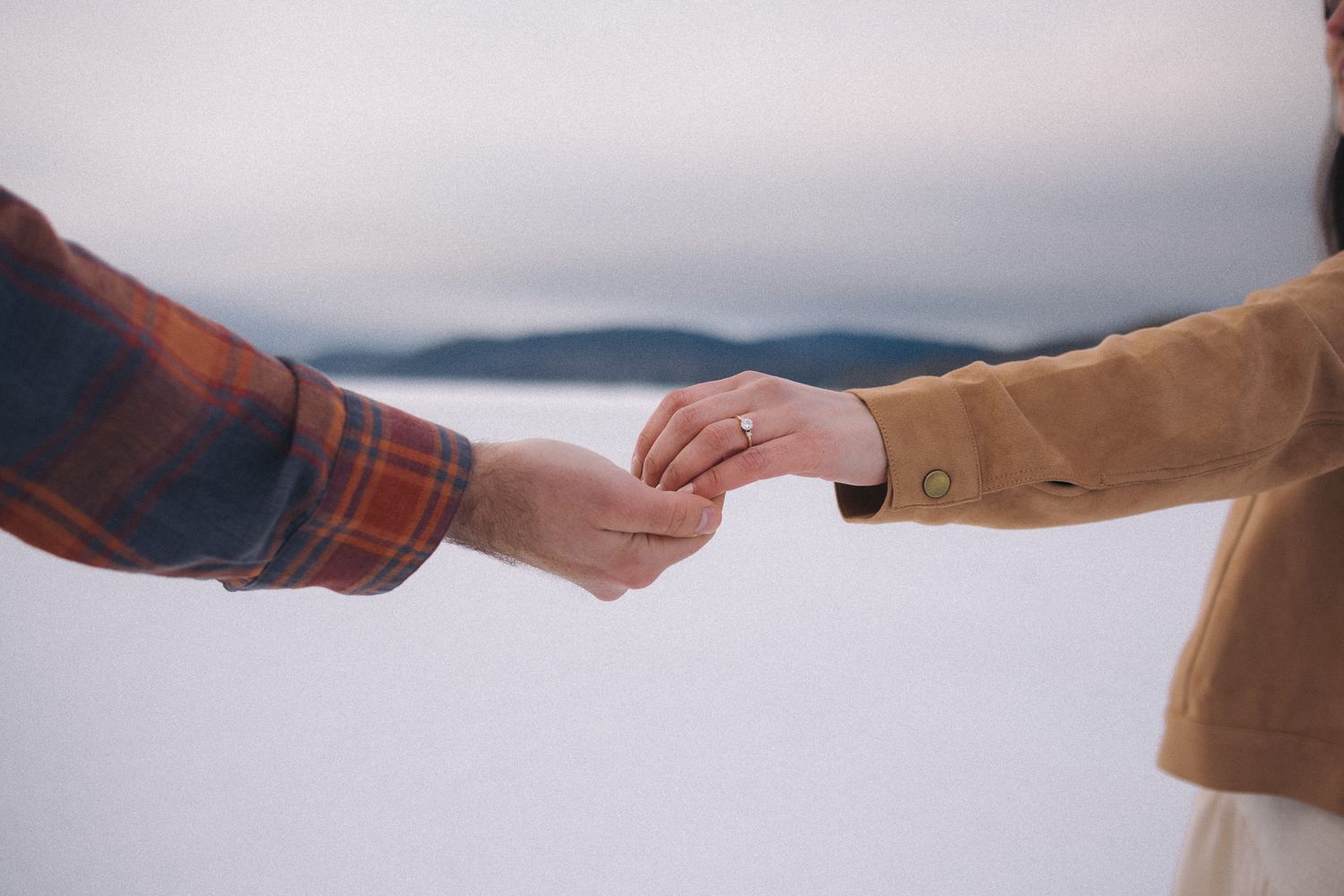 Two people gently holding hands outdoors in a snowy winter landscape, one wearing a ring.