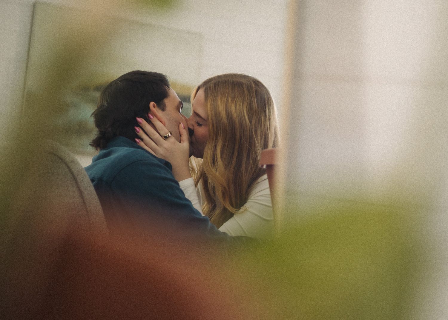 A couple shares a romantic kiss indoors, captured through soft, blurred green foliage in the foreground.