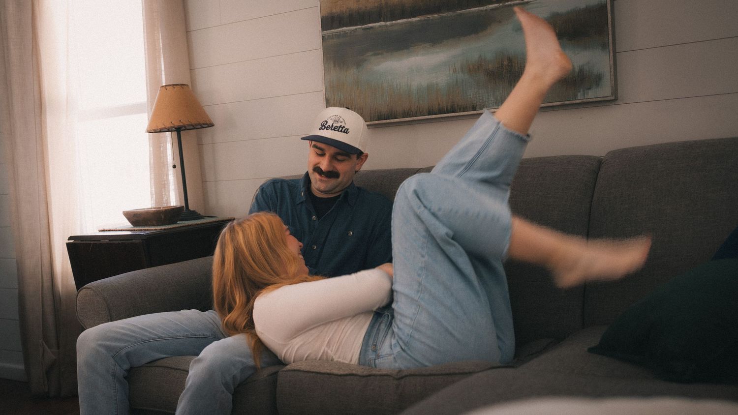 A couple laughing together on a gray couch in a cozy living room, woman resting legs up playfully.