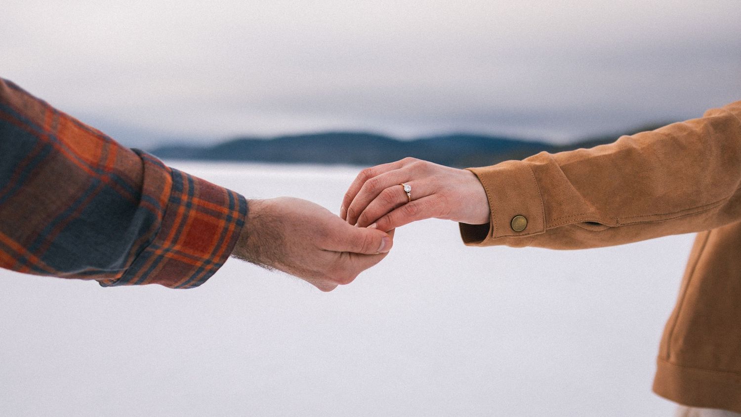 Two people gently holding hands outdoors, one wearing a ring, with a misty lake and mountains in the background.