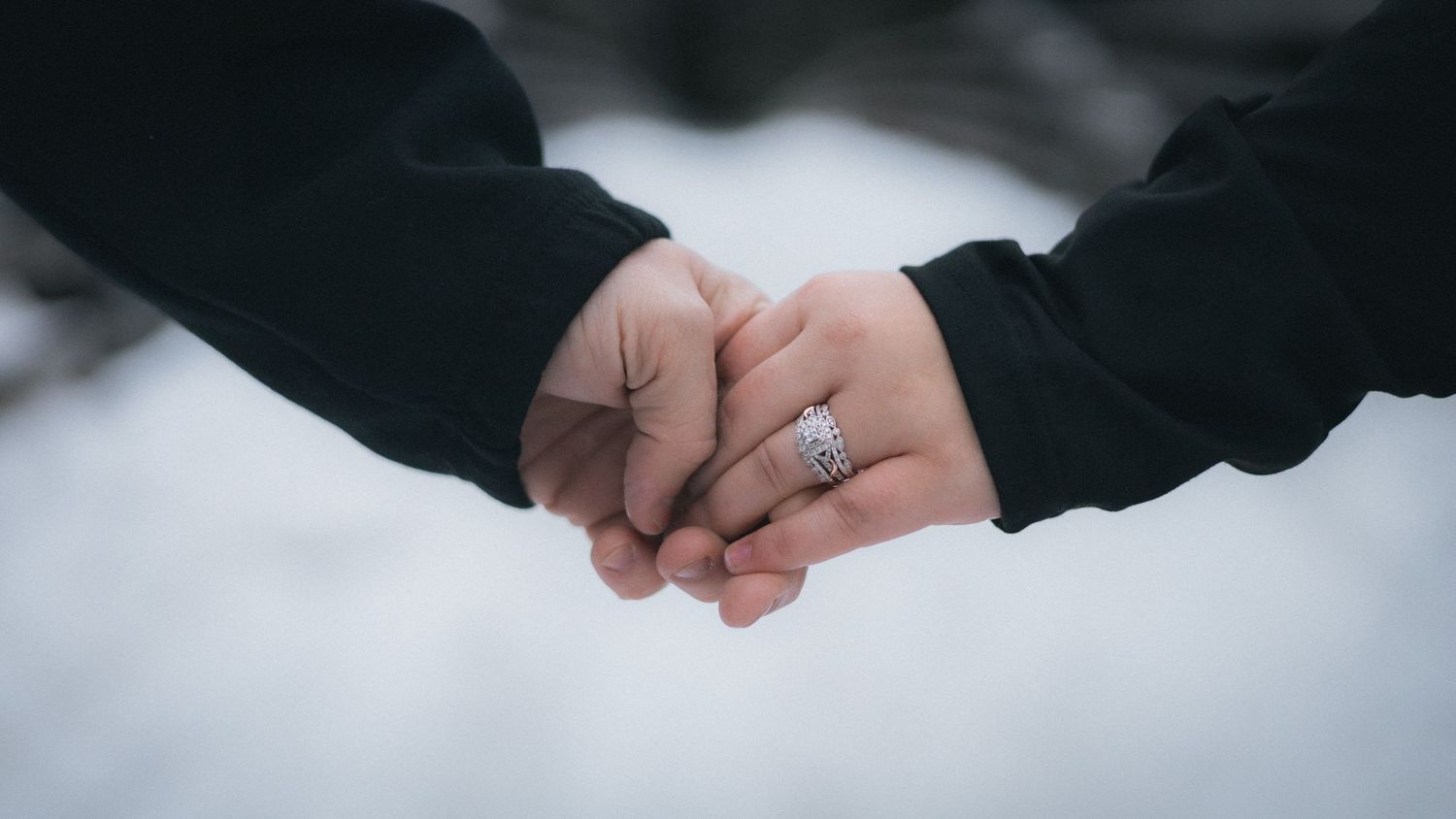 Two people hold hands while wearing dark clothing against a snowy white background, with an engagement ring visible.
