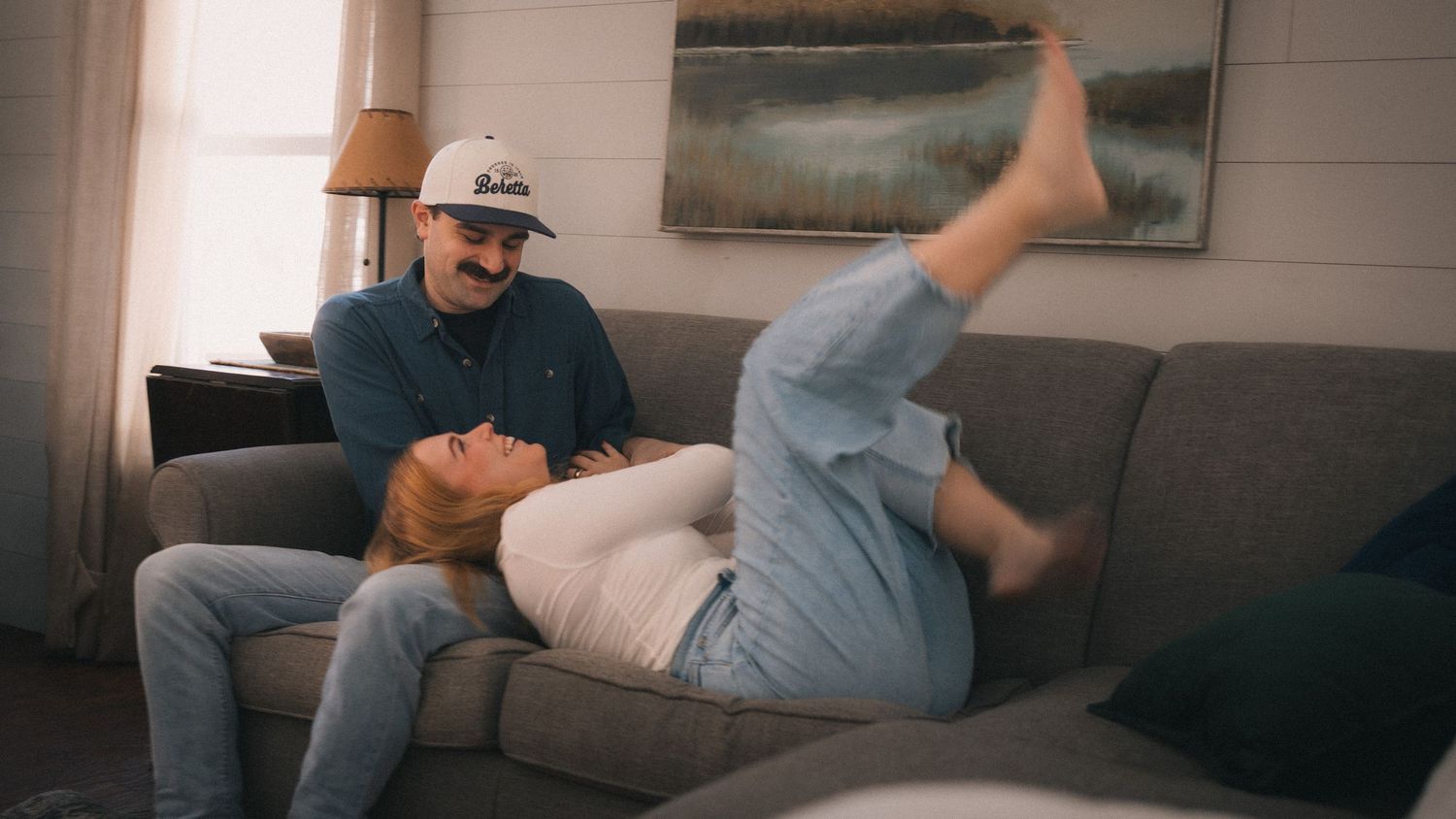A person relaxing on a grey couch in a living room while wearing casual attire and a baseball cap.