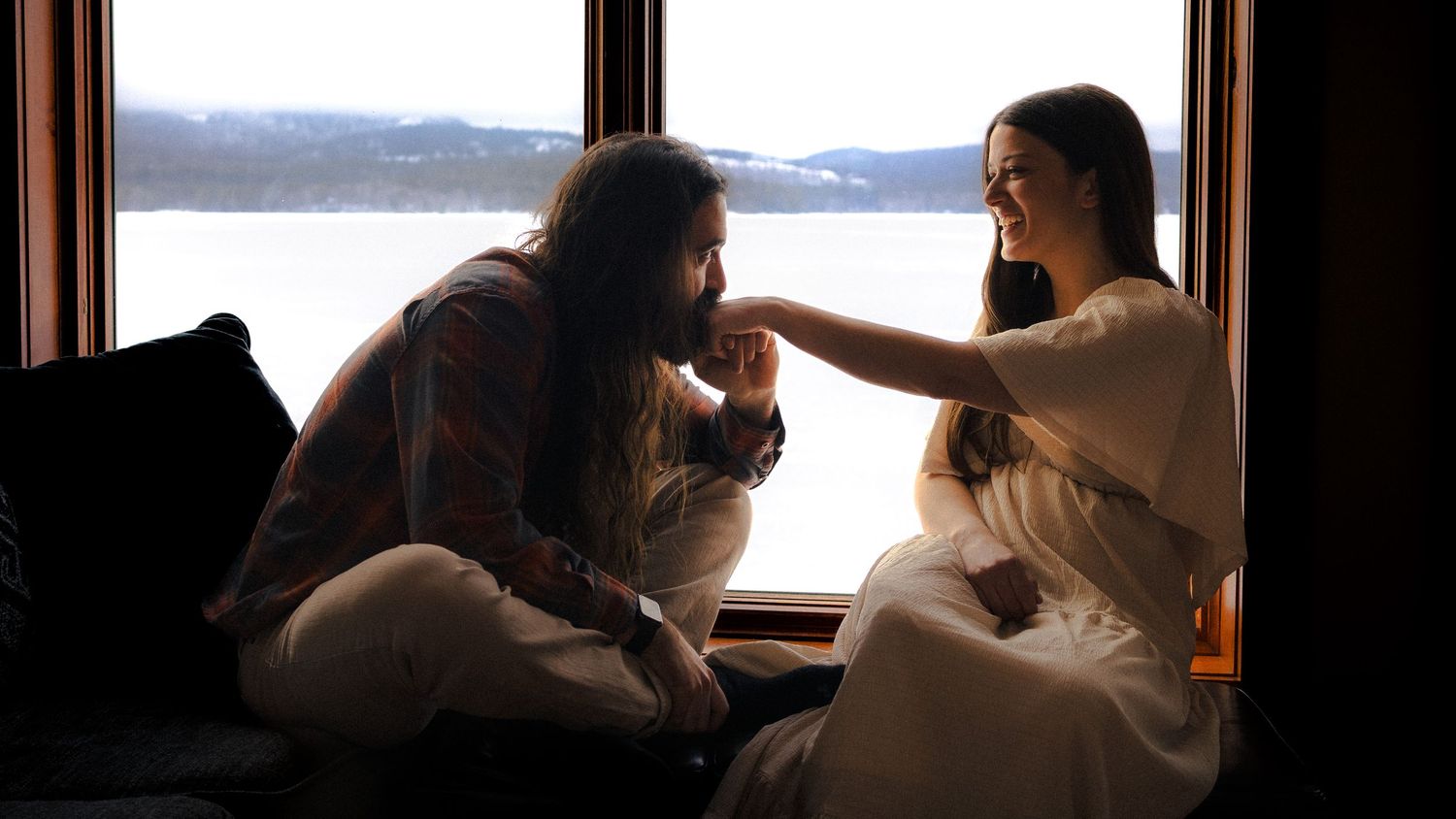Two women share an intimate conversation by a large window with a scenic lake view in a cozy, dimly lit room.
