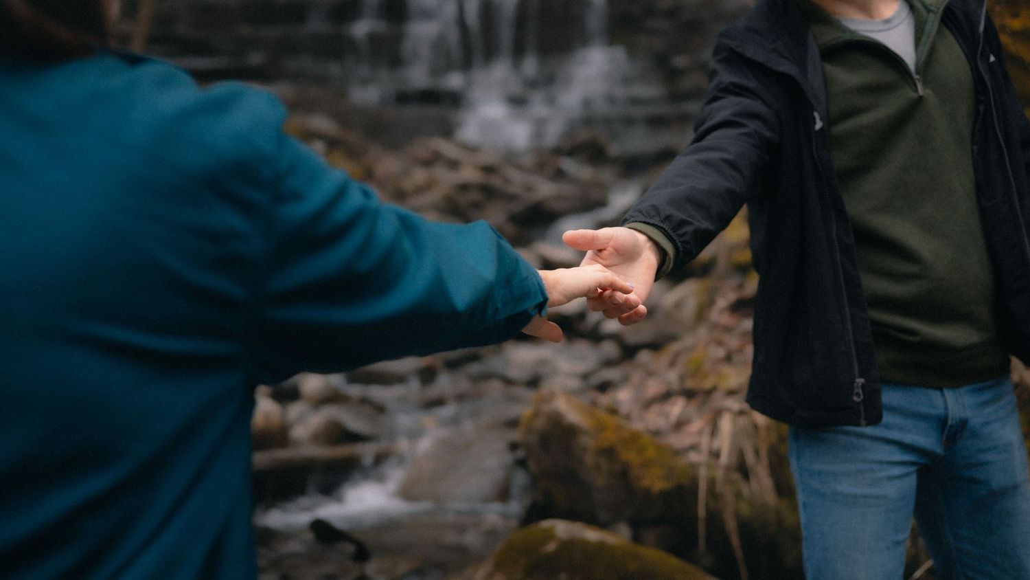 Two people holding hands near a rocky waterfall in a tender outdoor moment.