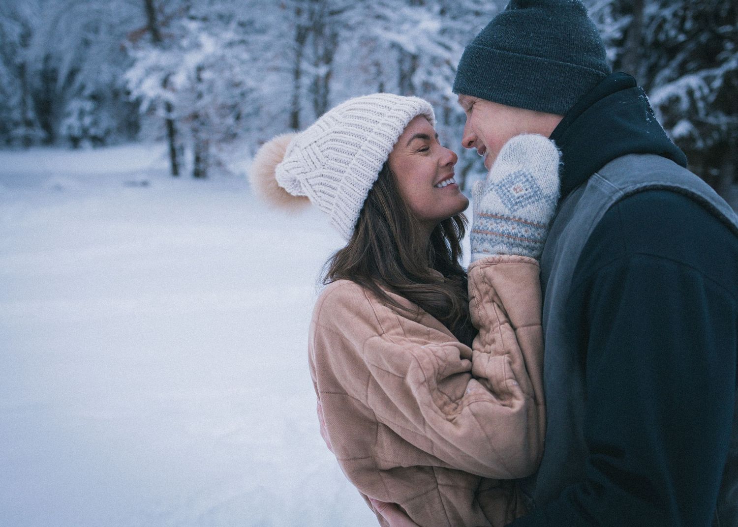 A couple in winter clothing shares a romantic moment in a snowy forest setting.