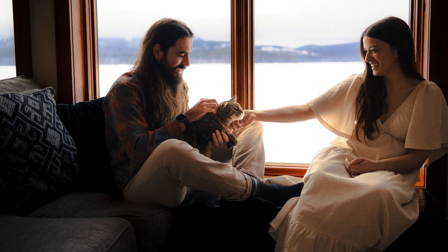 A couple sits together by a window looking out at a scenic mountain lake view at sunset.