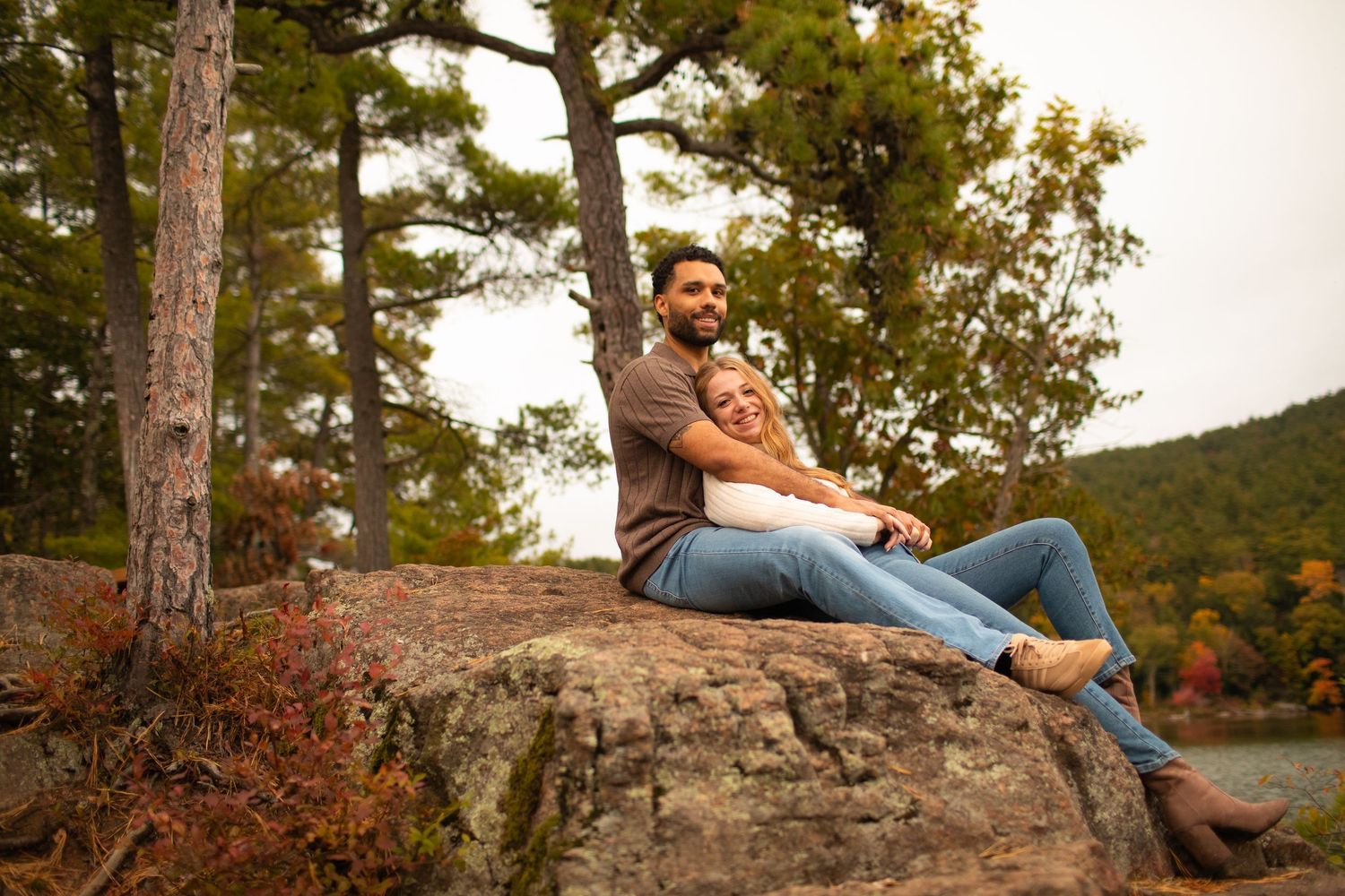 Couple sitting on a large rock surrounded by autumn trees near a lake.