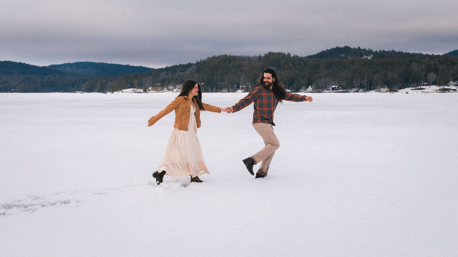 Couple holding hands dancing on a frozen snowy lake in winter, wearing bohemian wedding attire with pine trees behind.