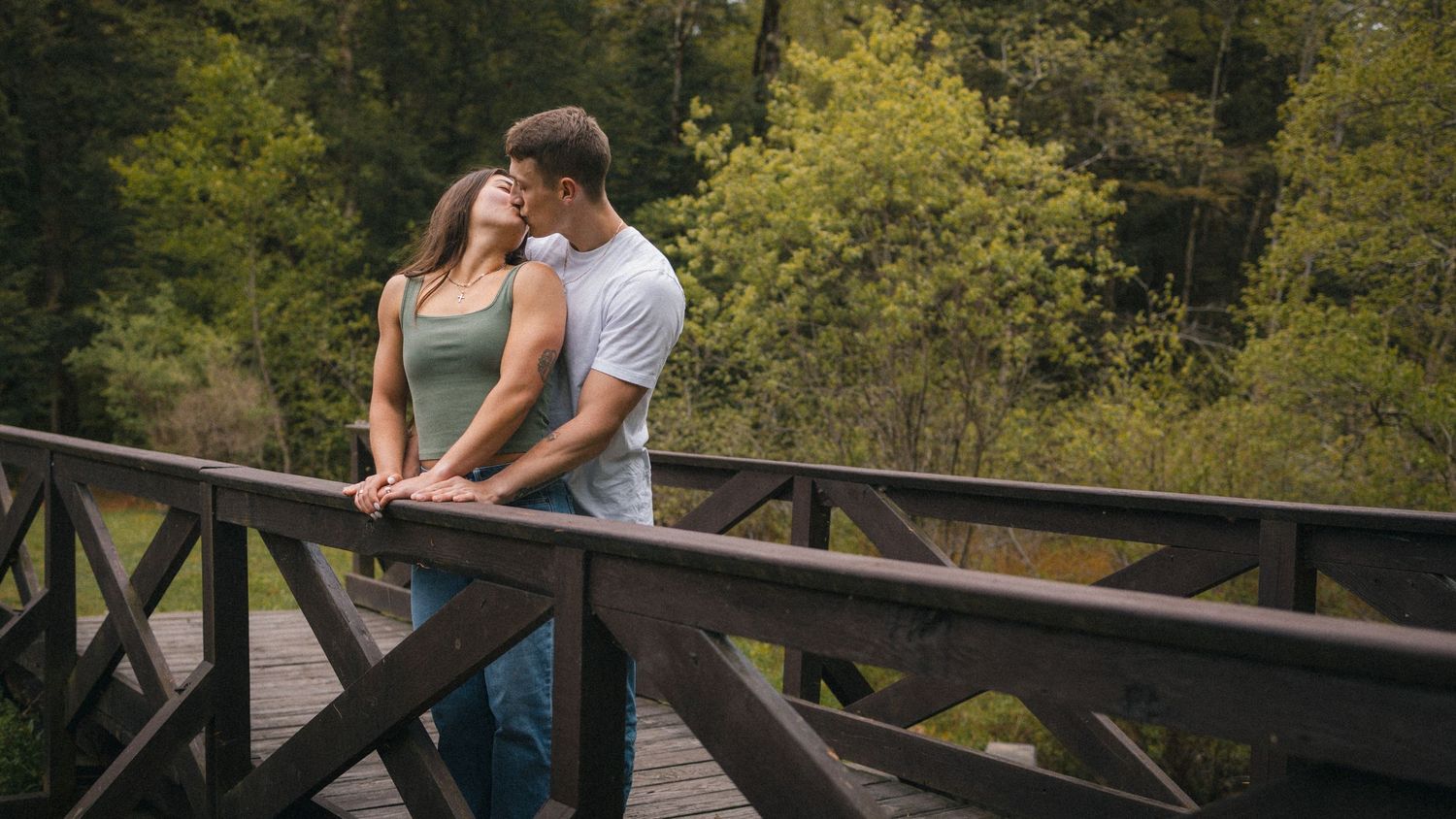 Romantic couple sharing a tender moment on a wooden bridge surrounded by lush green forest trees.