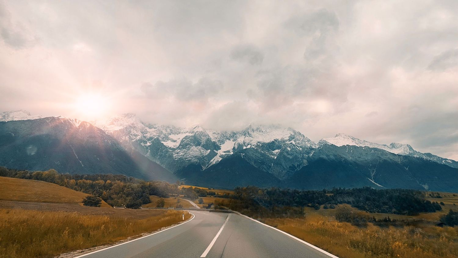 Panoramic mountain landscape with snow-capped peaks and winding road at sunset.