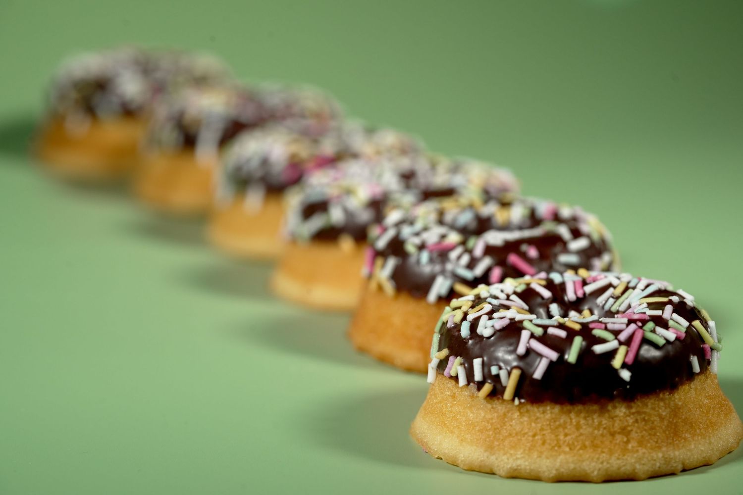 Row of chocolate-glazed mini donuts with white sprinkles on a green background.