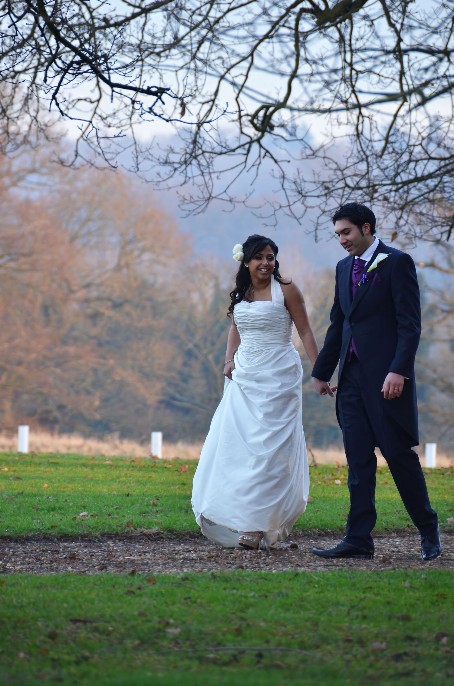 Newlywed couple walking through an autumn park with bare trees in the background.