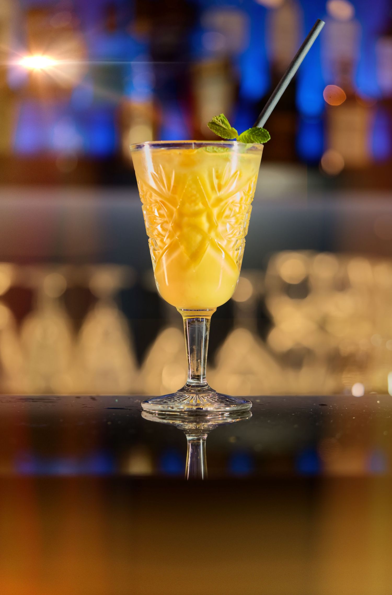 A golden cocktail served in an ornate vintage glass with garnish sits on a dark bar counter with blurred background lighting.
