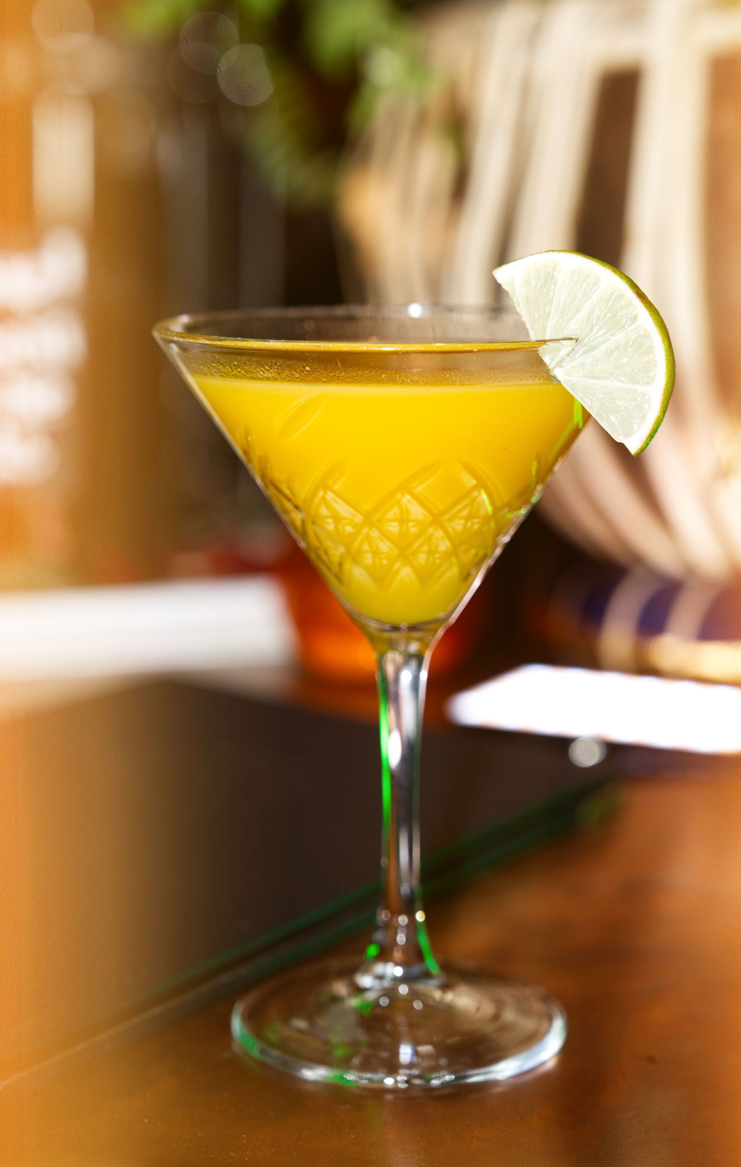 A golden yellow cocktail served in a martini glass with a lime garnish on a wooden bar surface.