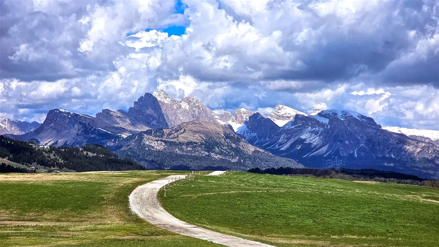 Panoramic mountain landscape with winding road through green meadows and snow-capped peaks.