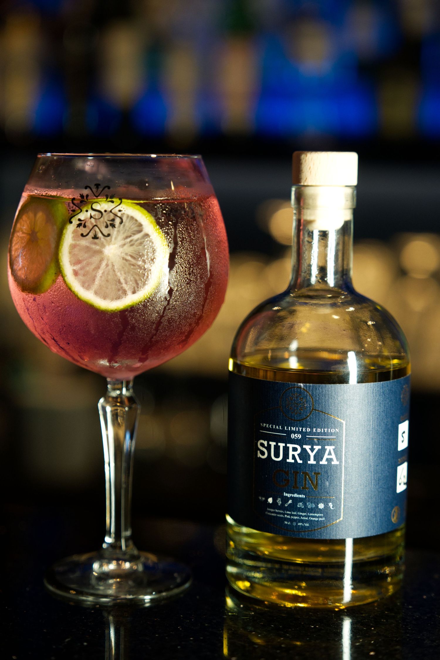 A red cocktail garnished with lemon slice sits next to a dark bottle of Suria liquor in a dimly lit bar setting.