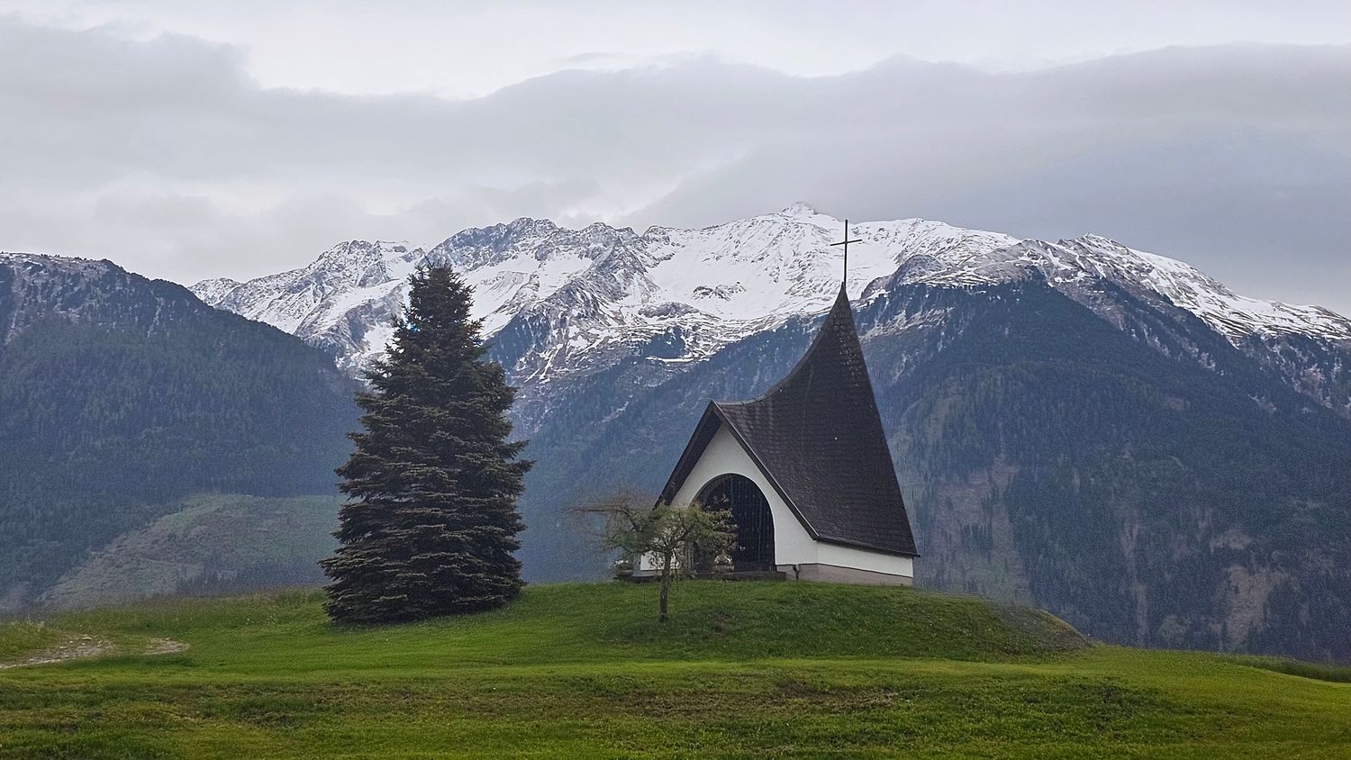 Panoramic mountain landscape with snow-capped peaks and evergreen trees against a cloudy sky.