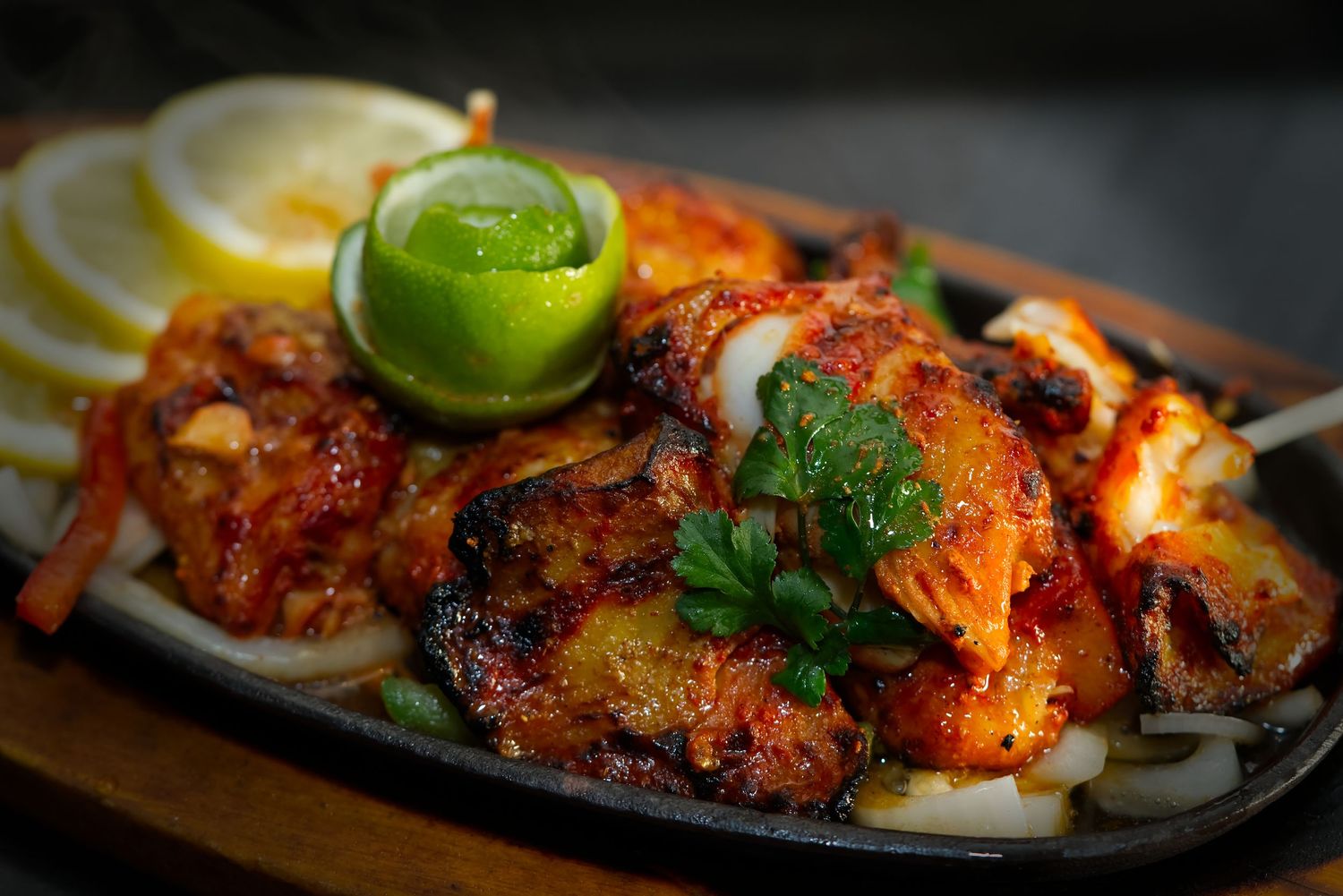 Sizzling grilled tandoori chicken pieces garnished with lime wedges and fresh herbs on a metal serving platter.