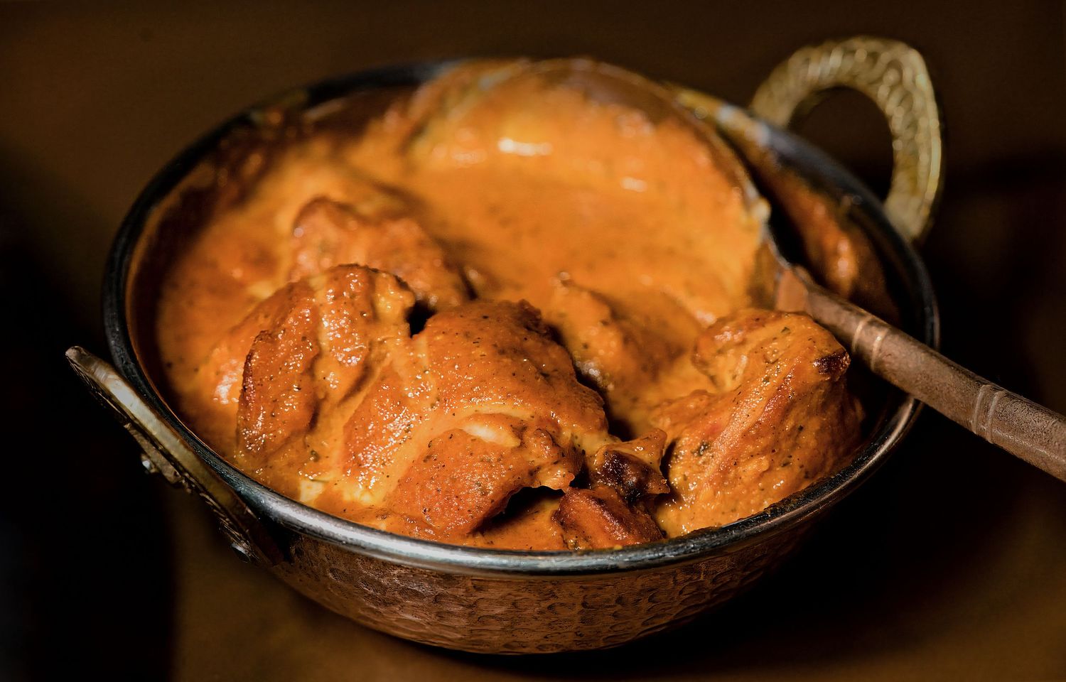 A rich and creamy butter chicken curry served in a traditional copper karahi bowl with a wooden spoon.