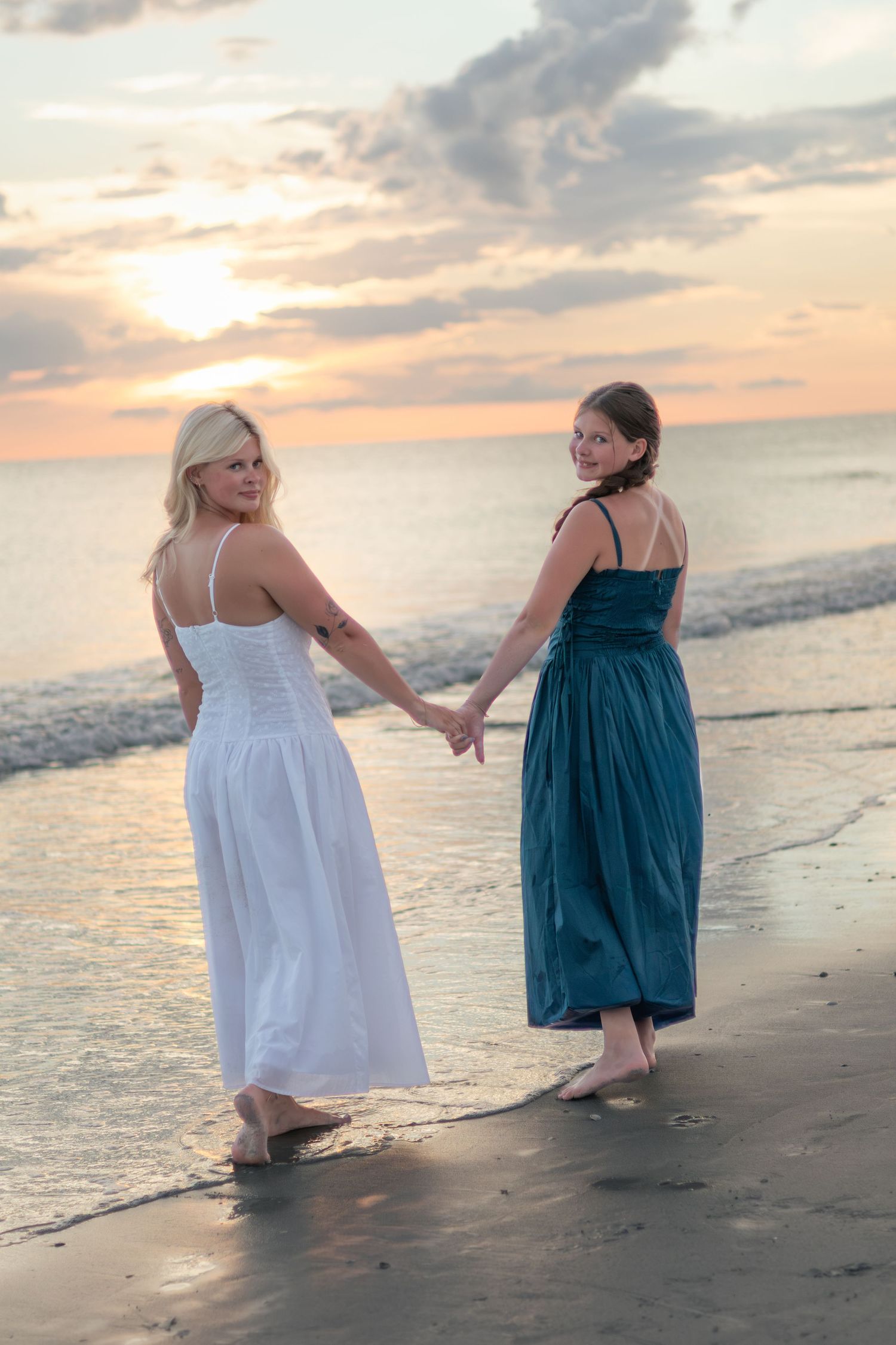 Two women hold hands while walking along the shoreline at sunset, one in a white dress and the other in a blue dress.