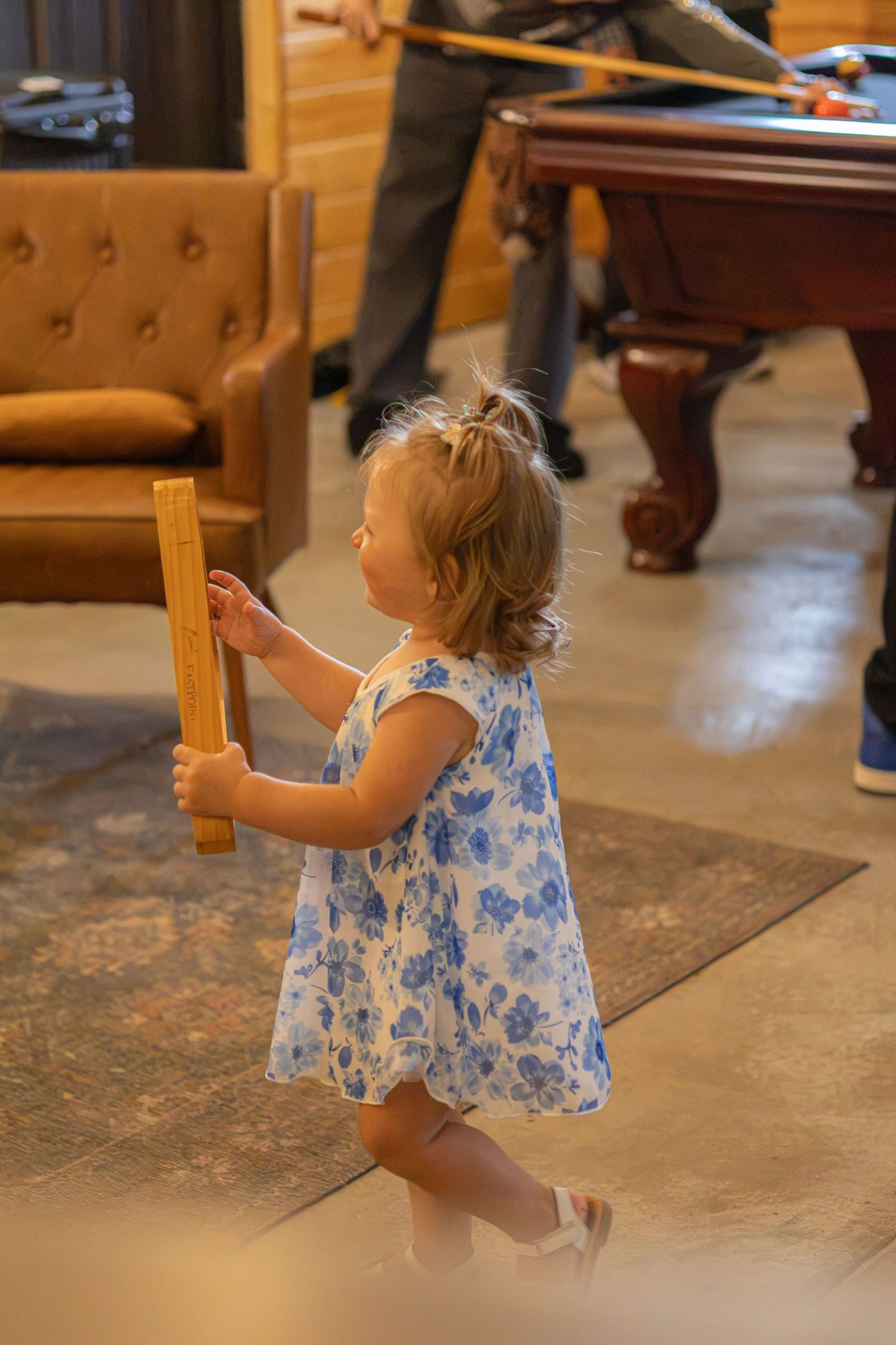 Toddler girl in a blue floral dress carries a wooden block across a rug in a room with a pool table.