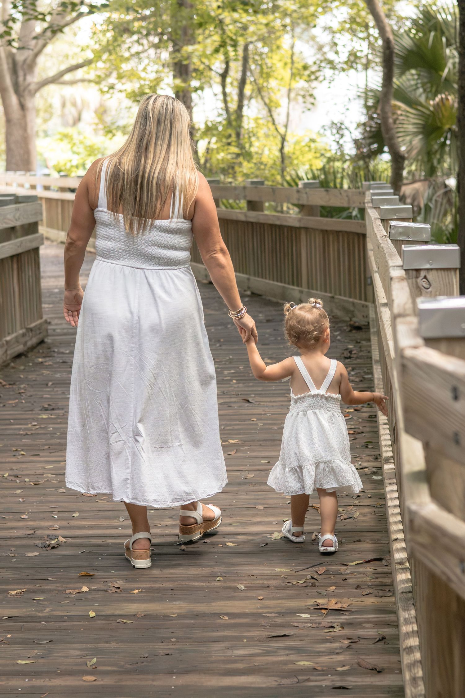 A woman and a toddler in matching white dresses walk hand in hand along a wooden boardwalk lined with trees.