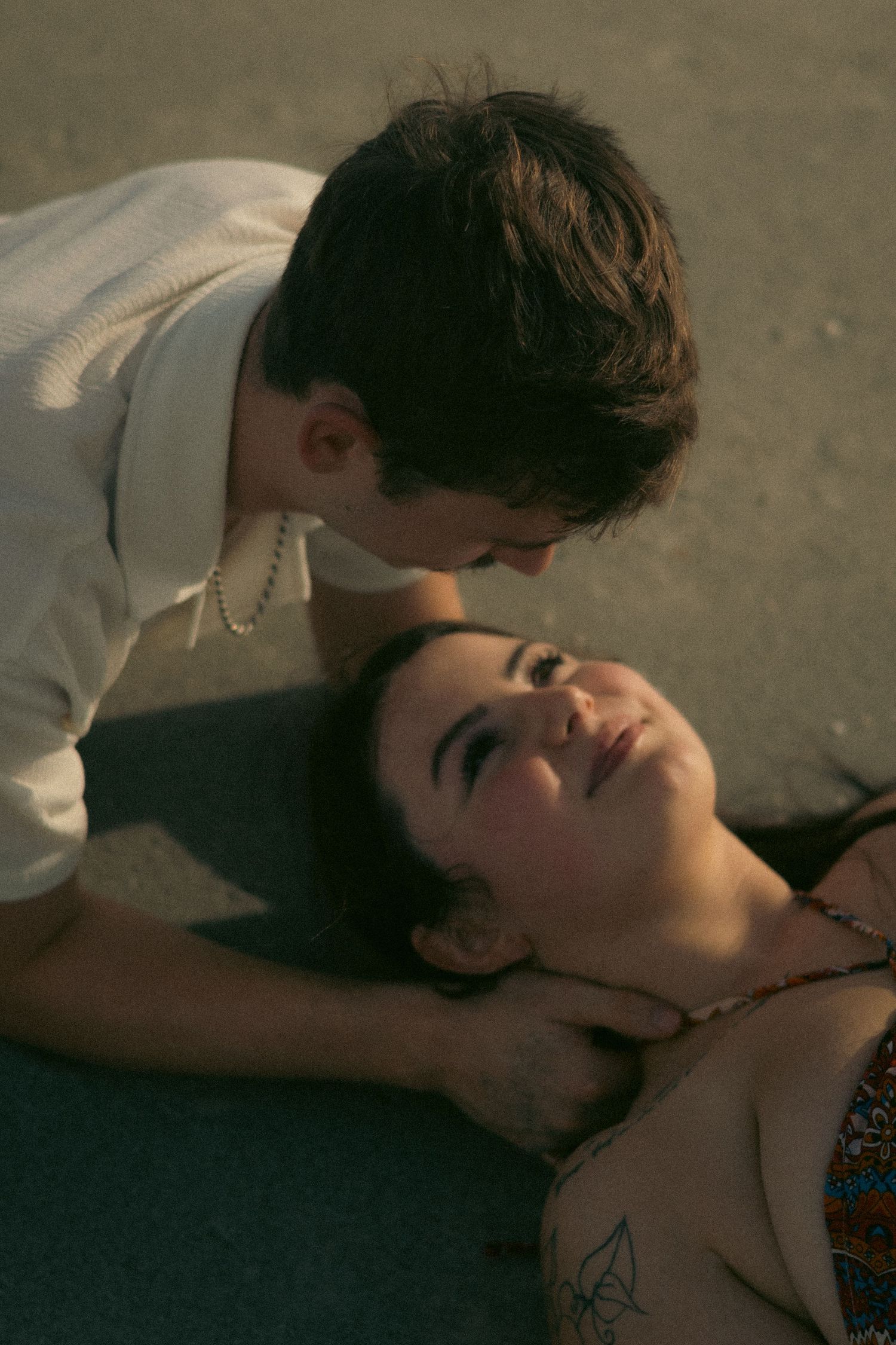 Overhead view of a couple in warm golden light, a man leaning over a smiling woman with a tattoo as she lies on the ground.