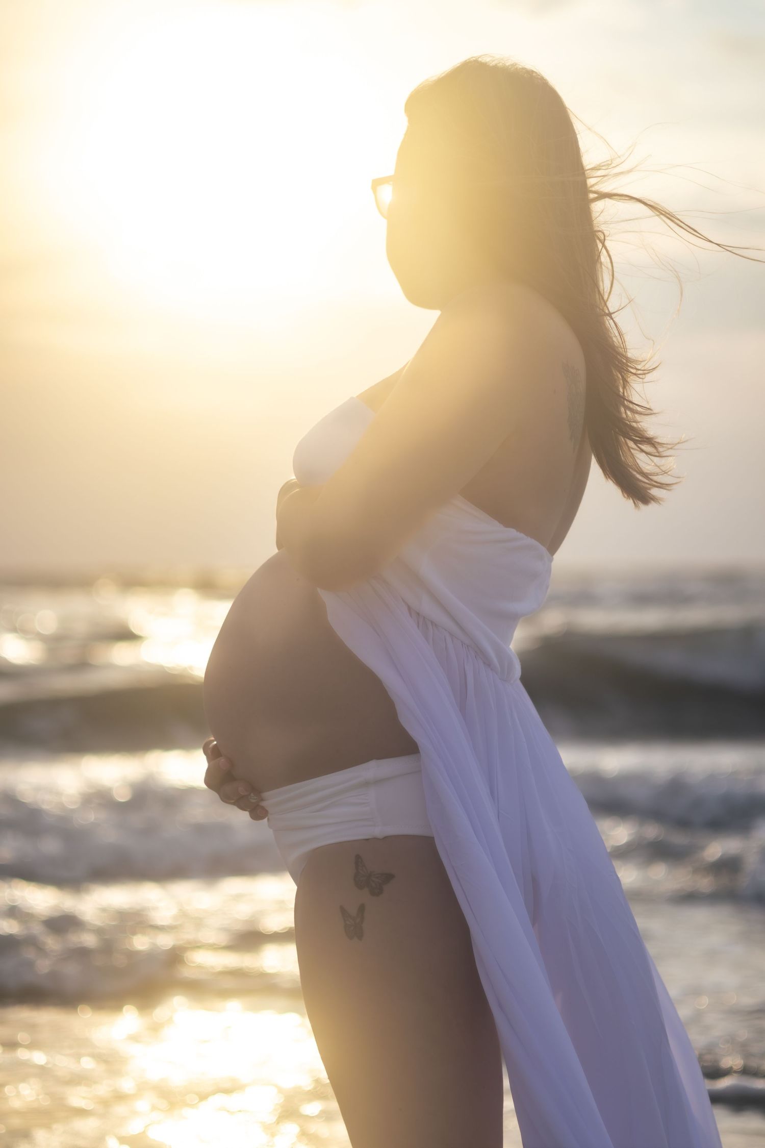 Silhouette of a pregnant woman in white holding her belly against a golden sunset over sparkling ocean waves.