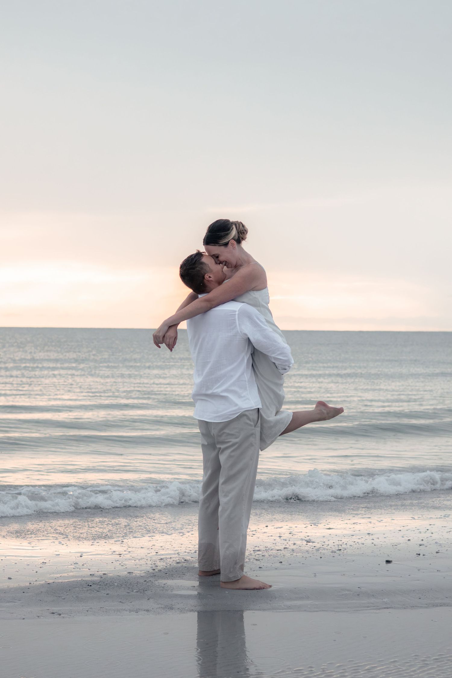 Barefoot man lifts his smiling partner with her leg kicked up as they kiss on the shore of a calm beach at sunset.