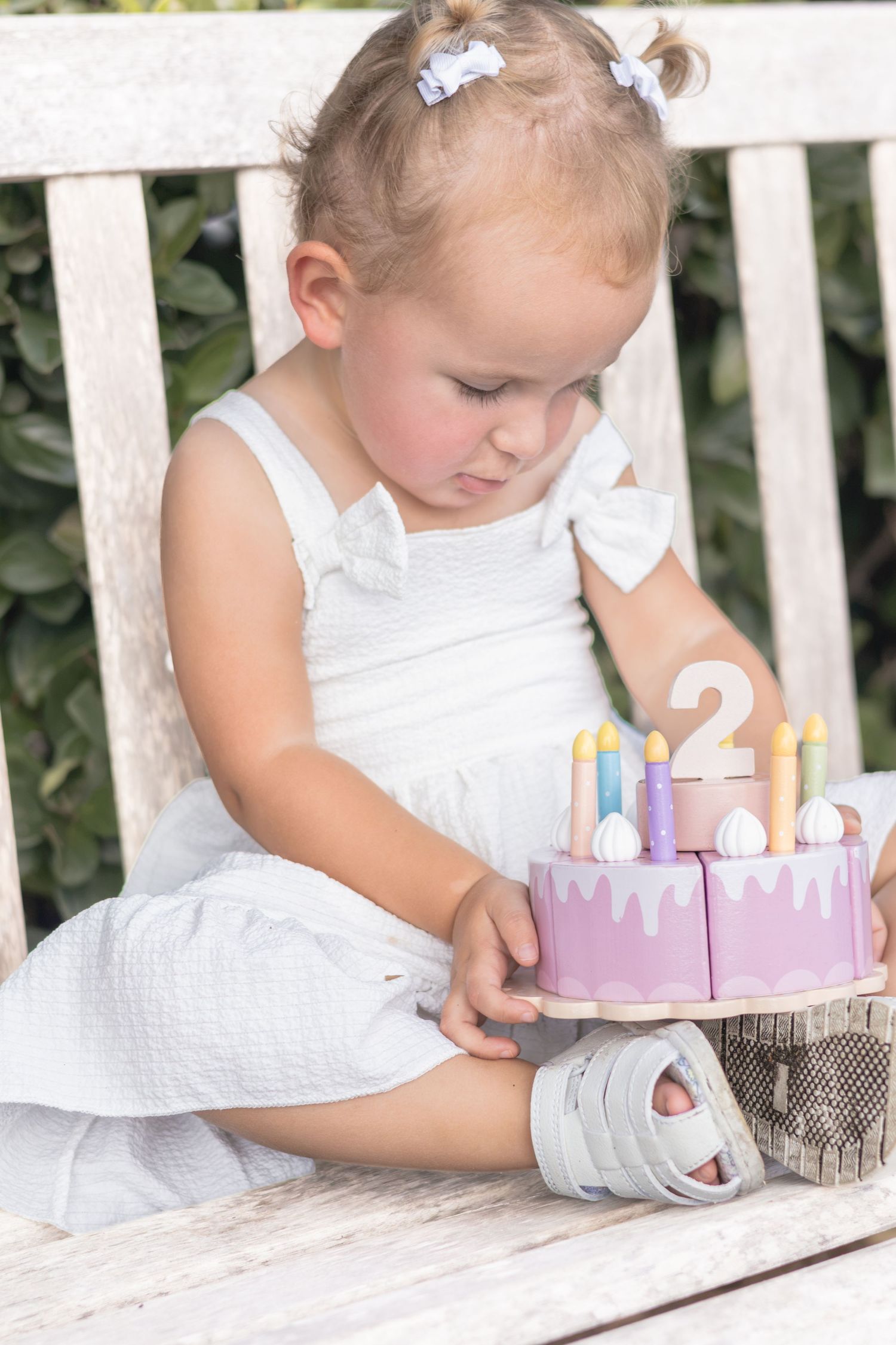 Toddler in a white dress and hair bows sits on a wooden bench, looking at a pink wooden toy birthday cake with candles and a number two.