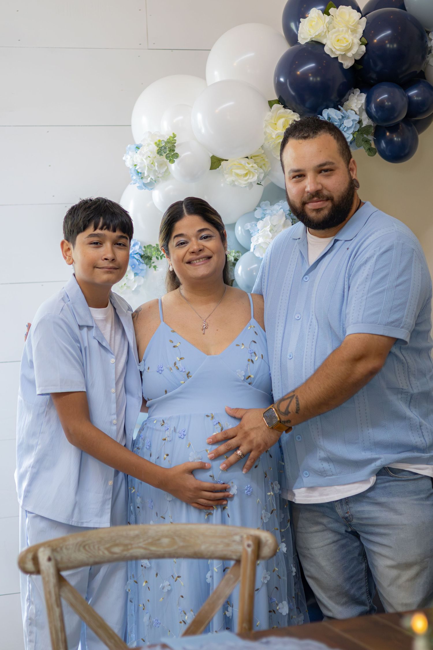 A smiling family of three in matching light blue outfits poses beside a blue and white balloon arch at a baby shower celebration.