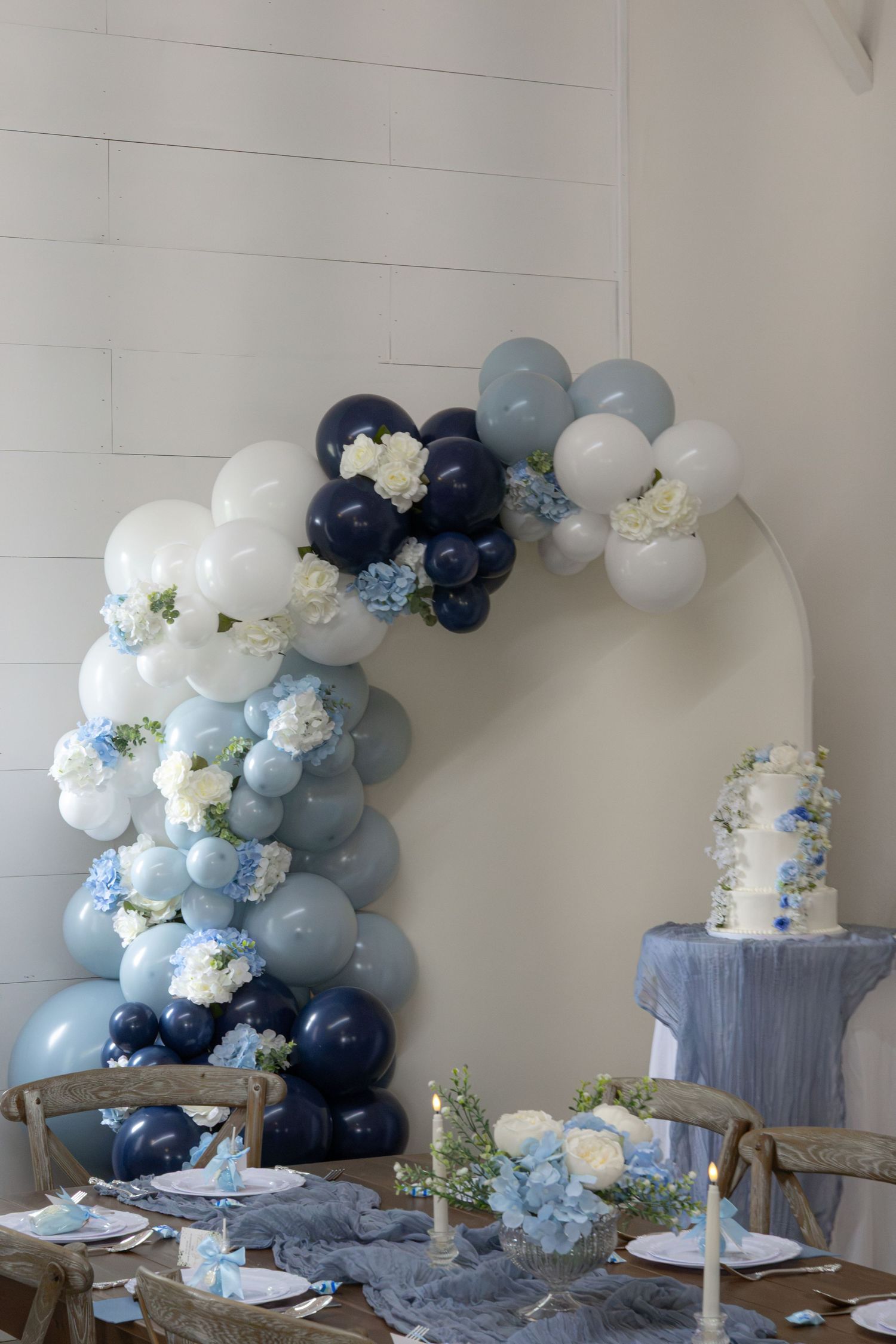 Blue and white balloon arch with fresh flowers above a rustic wooden table set for a baby shower, with a floral cake nearby.