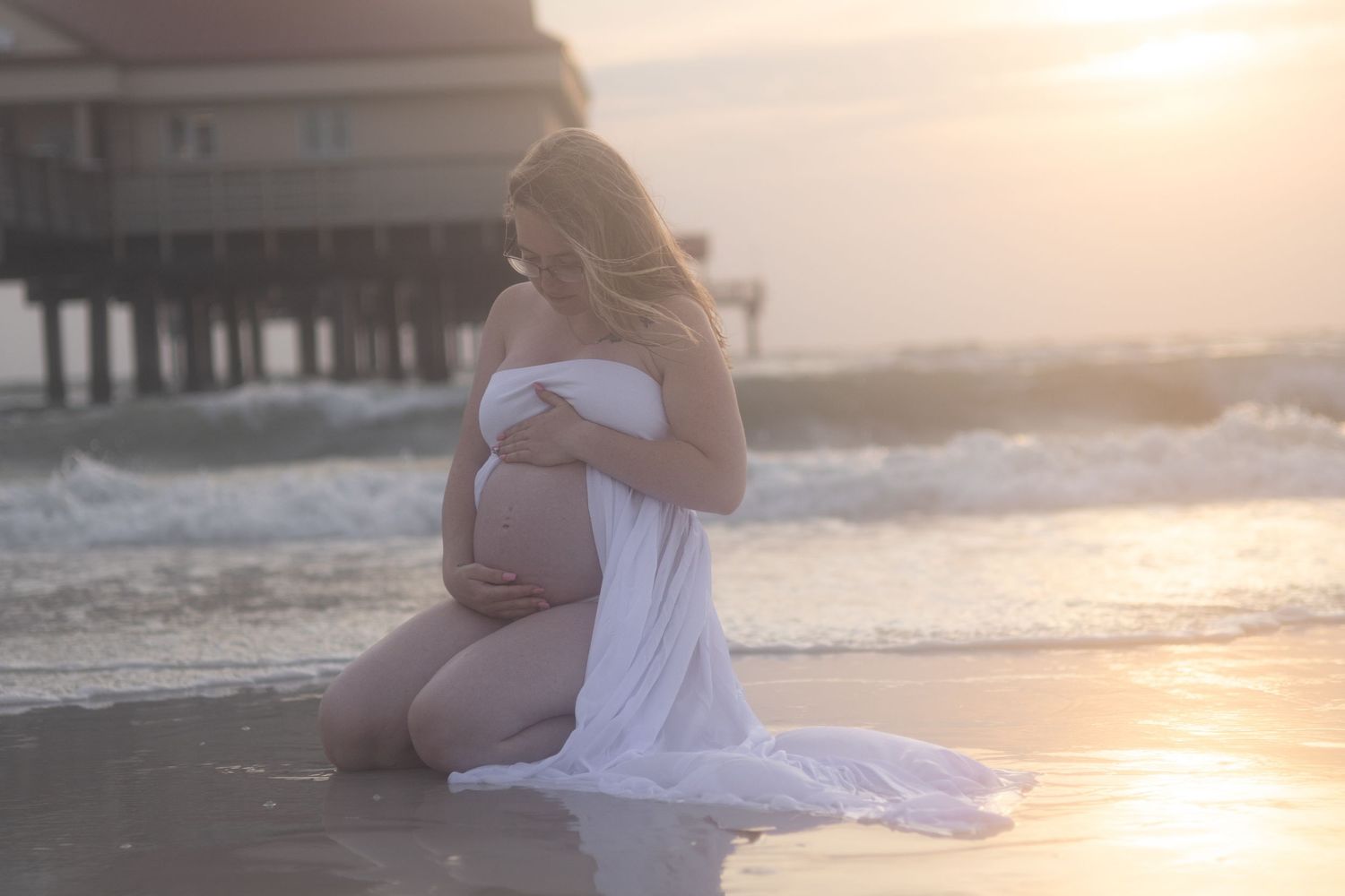 Pregnant woman in a flowing white wrap kneels on the wet sand at sunset with a pier and ocean waves behind her.