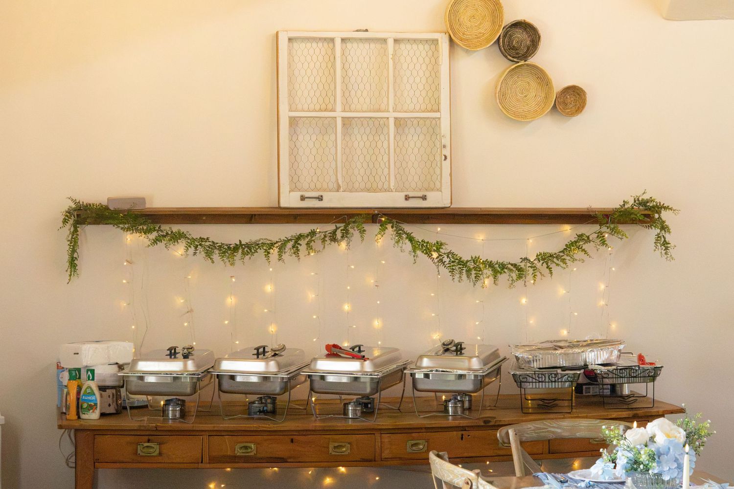 Buffet setup with chafing dishes on a wooden table beneath a greenery garland and string lights, with woven baskets on the wall.