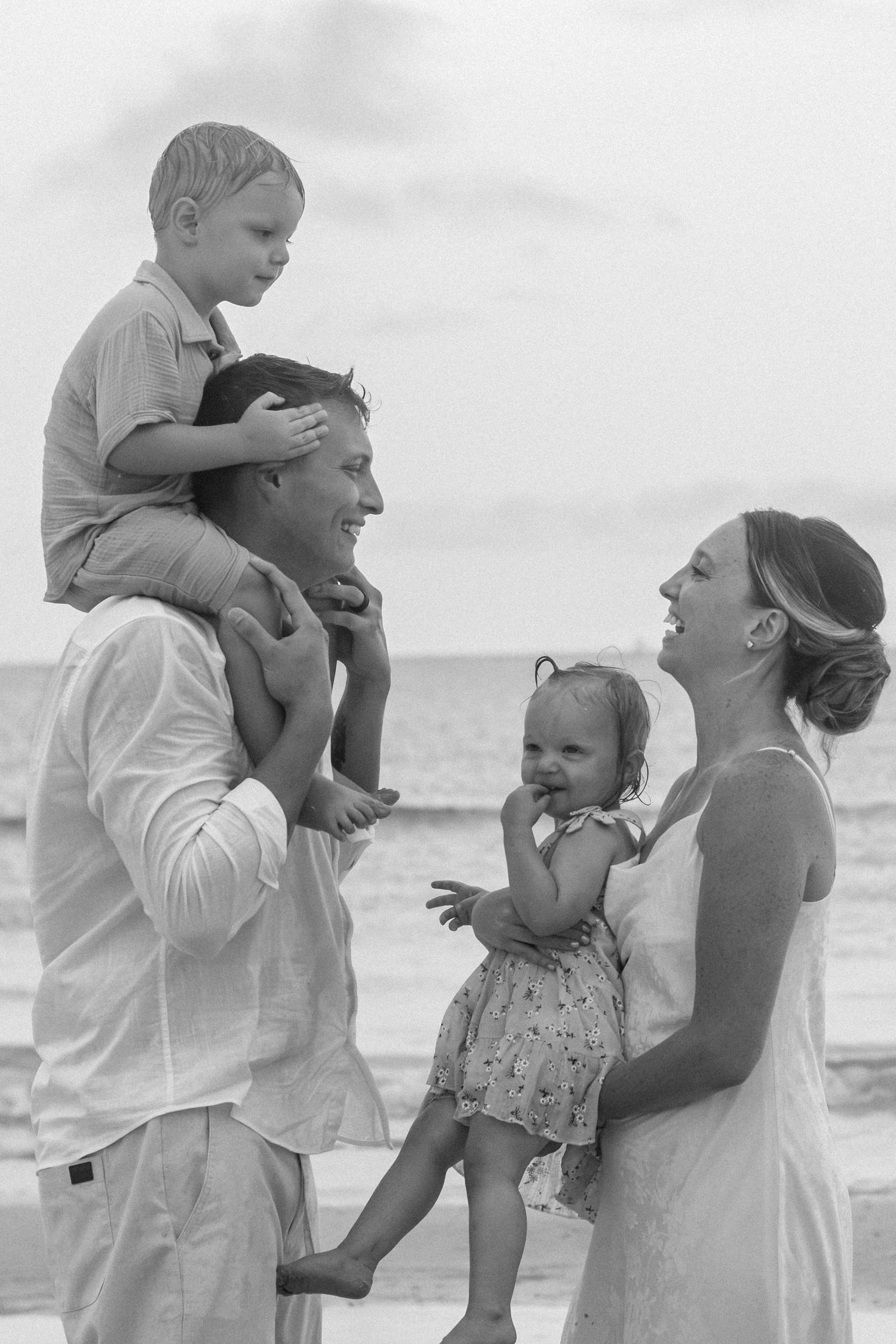 Black and white photo of a laughing family on the beach, with a boy on dad's shoulders and mom holding a toddler girl.