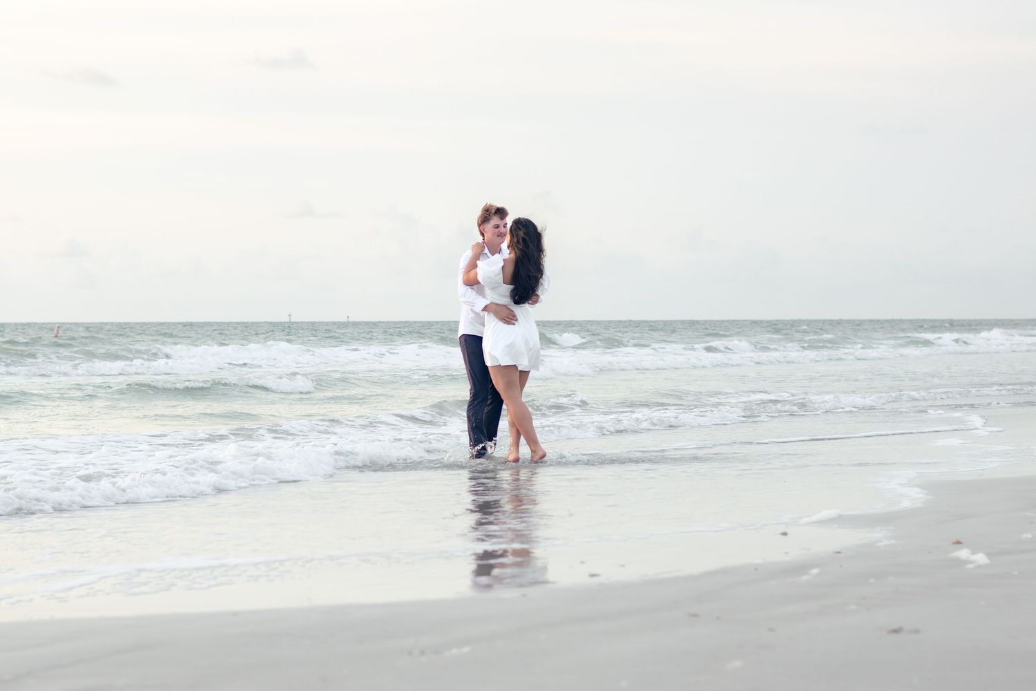 Couple in white embracing at the water's edge on a quiet beach with gentle waves rolling in.