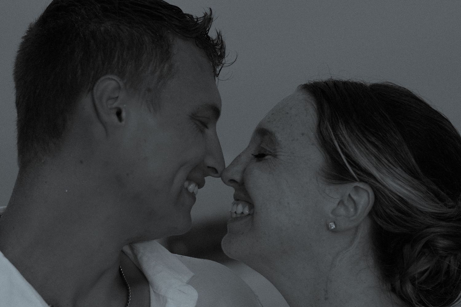 Smiling couple standing nose to nose in a tender black and white portrait, sharing an intimate and joyful romantic moment together.